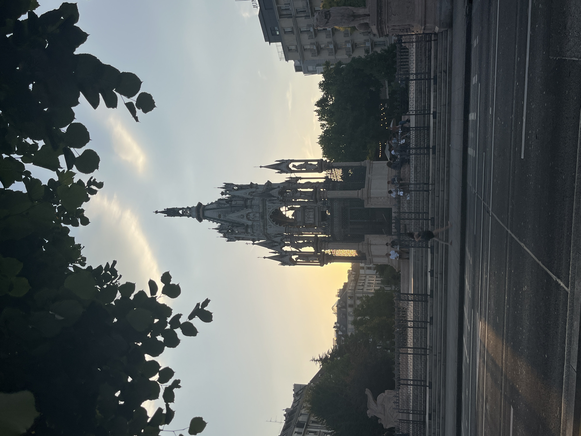 This is an outdoor, ground-level, eye-level shot of the Brunswick Monument in Geneva, Switzerland, at sunset. The monument is a Gothic-style mausoleum with intricate carvings and spires, surrounded by a fence. Several people are seated on steps in front of the monument, while one person walks on a sidewalk in the foreground. Buildings, trees, and a street with traffic lanes are also visible. The sky is a pale yellow and blue, with clouds and the setting sun casting a warm glow over the scene.