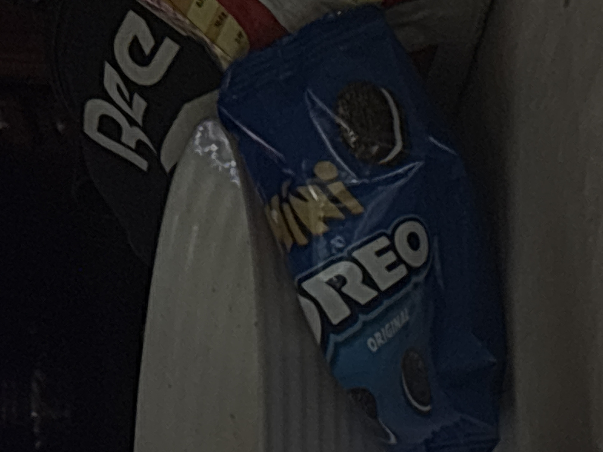 This image captures an indoor scene in the city of Burgas, Bulgaria, presenting a close-up view of several objects on a flat surface. The central object is a small, blue plastic packet of "Mini OREO Original" cookies, positioned diagonally across the lower-middle portion of the frame. The word "MINI" is printed in gold lettering above "OREO," which is in white capital letters. A graphic of a chocolate sandwich cookie with white cream filling is visible on the right side of the packet.
To the left of the Oreo packet, a portion of a light-colored, possibly off-white or light grey, bowl or dish is visible, its rim curving towards the top left. Behind and to the right of the Oreo packet, a dark fabric object, potentially a cap or piece of clothing, displays a stylized word in light grey capital letters, resembling "Bee" or "Ree." Further right, partially obscured, is an item with a grid pattern composed of small, rectangular sections in yellowish-brown tones, accompanied by a small visible section of red material. All objects rest on a flat, dark grey surface, likely a table. The scene is dimly lit, contributing to a muted color palette. No human figures or active interactions are depicted.