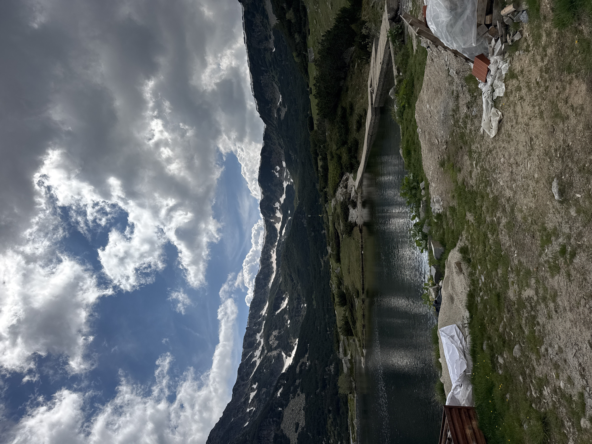 The image depicts a scenic mountain lake landscape in Avramovo, Bulgaria. The foreground shows a gravelly area with some construction debris, including a white tarp, a rusty metal structure, and piles of sand and bricks. A dark, still lake occupies the middle ground, reflecting the sky and surrounding mountains. The far background is dominated by a majestic mountain range, with dark green pine forests covering its lower slopes and patches of snow visible on the higher elevations. The sky is a dramatic mix of bright blue and heavy, white and grey clouds, suggesting dynamic weather. The scene is natural and serene, with a touch of human presence in the form of the construction materials in the foreground.