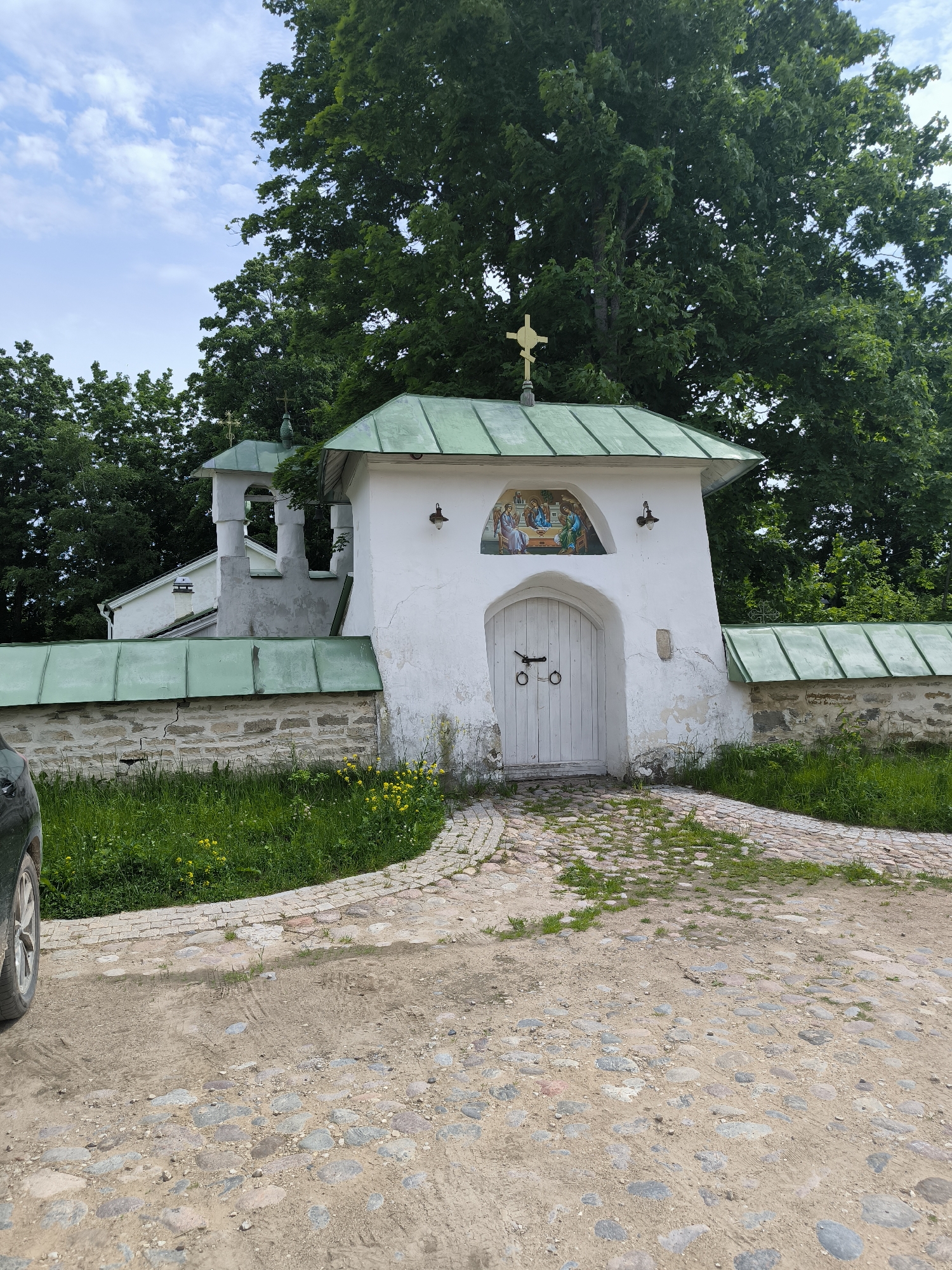 The image shows a white arched gateway with a double wooden door, decorated with a religious icon above it. The gateway is part of a stone wall topped with a green metal roof. Behind the gateway, a bell tower with a similar green roof is visible, attached to a white building. Lush green trees form a backdrop against a partly cloudy sky. The ground in front of the gateway is paved with cobblestones, leading from a dirt path. The scene is located in Pechory, Russia, possibly at the entrance to a monastery or church complex, given the religious iconography and architecture. The time of day appears to be daytime, with soft lighting.