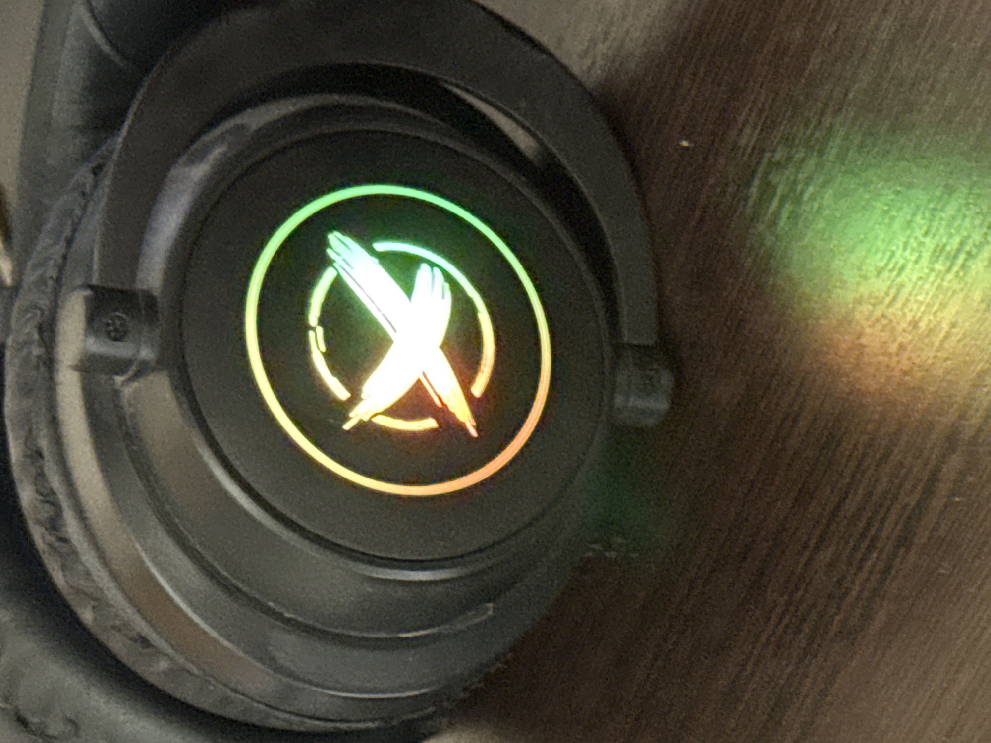 A close-up, eye-level shot shows a black gaming headset with a circular logo illuminated by a rainbow gradient of light, transitioning from orange to green. The logo features a stylized white "X" shape with pointed ends. The headset is resting on a dark brown wooden surface, which is reflecting the colorful light from the logo. The background is dark and out of focus, emphasizing the headset and its illuminated emblem. The lighting is low, with the primary light source appearing to be the headset's logo. The image is a static, indoor scene with no people or discernible actions.