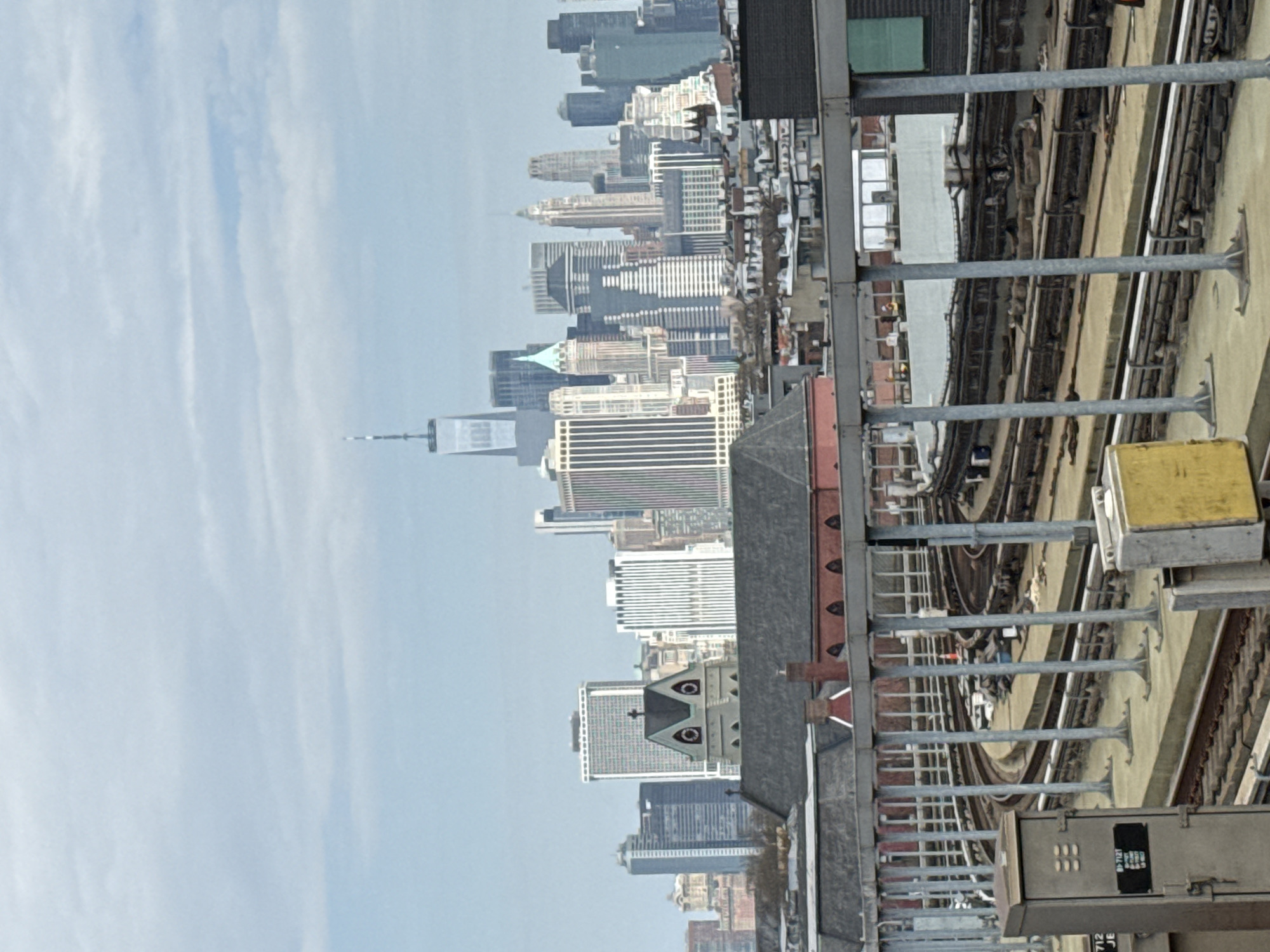 The image shows the New York City skyline from an elevated vantage point, likely near a subway line. Dominating the skyline is One World Trade Center. The image also features an older building with distinctive architectural features like round window accents and a pitched roof, partially obscured by the elevated subway structure. The subway structure is prominent, with tracks, support beams, and signaling equipment visible. The sky is overcast with visible wispy clouds.