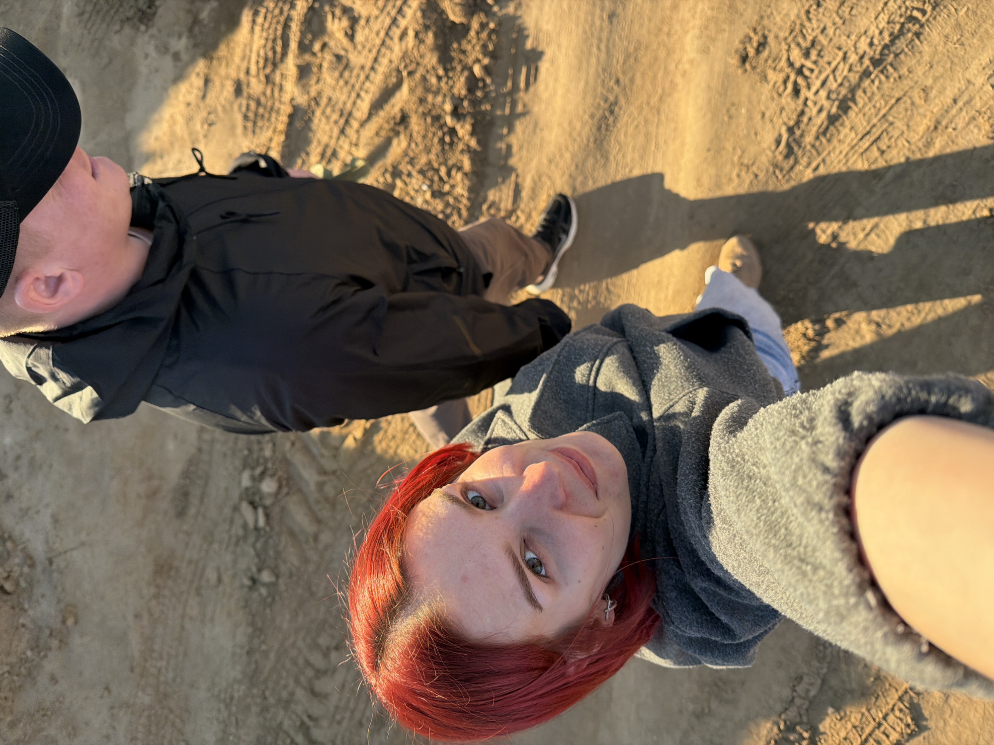 Two individuals are standing on what appears to be a dirt or sandy surface, possibly a path or road. The ground shows tire tracks and other markings. One person, presumably taking the photo, has red hair and is wearing a grey sweatshirt. The other person, partially visible, is wearing a black jacket, tan pants, and a black baseball cap. The scene is illuminated by sunlight, creating distinct shadows. The location is Chaikovsky, Russia, and the picture seems to be taken outside, with the ground surface suggesting an unpaved area.