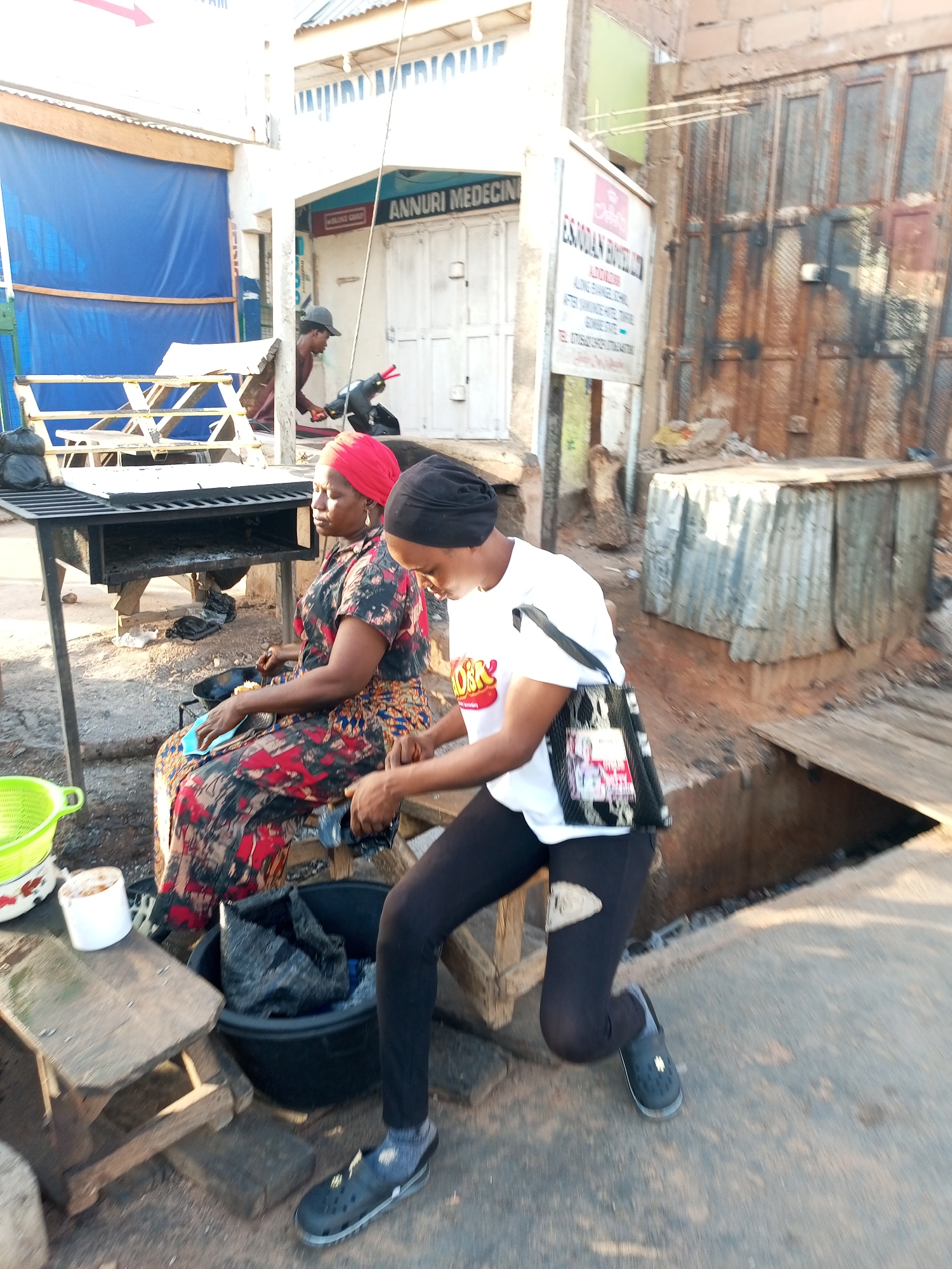 The image depicts an outdoor scene in Gombe, Nigeria, with two women engaged in an activity near a grill. The woman in the foreground, on the right, is wearing a black head covering and a white t-shirt with a graphic print, paired with black leggings and dark grey crocs. She is in motion, leaning forward with her right arm extended. To her left, another woman is seated, wearing a red headscarf and a patterned dress. She is looking down.
In the background, a metallic grill with charcoal is visible. Various objects are scattered around, including a green plastic bowl, a white mug, and a large black tub containing a black plastic bag. The environment suggests a market or roadside stall setting, with buildings and corrugated metal structures in the background. A sign with text is partially visible on a building. The ground is paved with concrete. The overall lighting indicates it is daytime.
