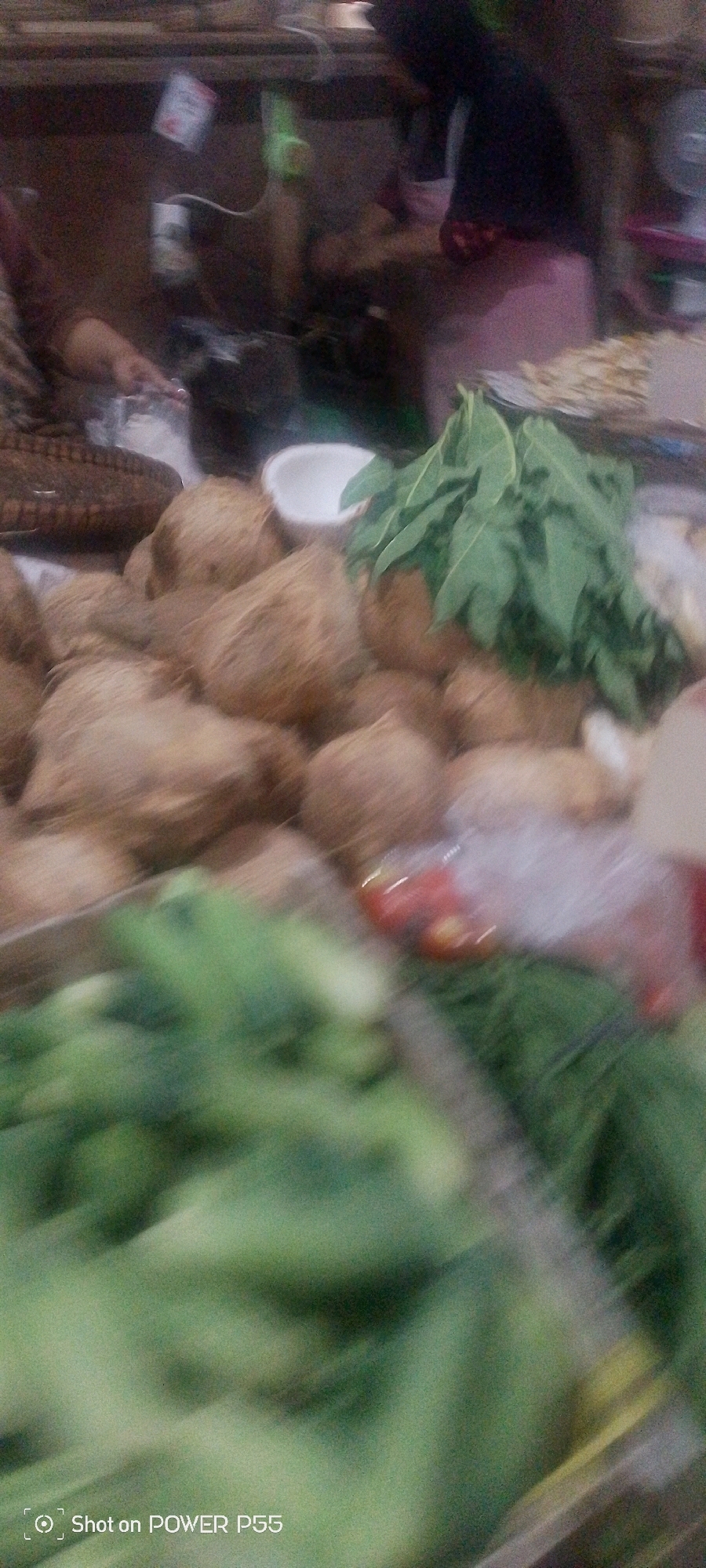 A low-angle, medium shot captures a blurry marketplace scene. The foreground is dominated by out-of-focus green vegetables. In the middle ground, a pile of coconuts is visible, with a bunch of leafy green vegetables and a white bowl placed amongst them. Behind the coconuts, a person wearing a dark garment and a pink apron is partially visible. The background shows more blurry figures and market stalls. The image has a watermark in the bottom left corner that reads "Shot on POWER P55".