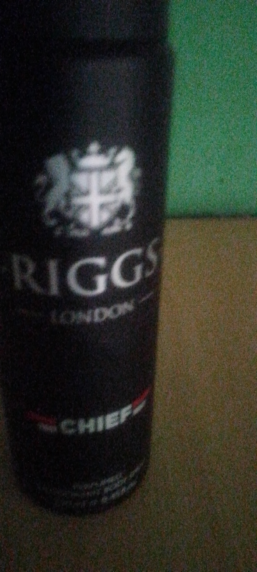 The image is a close-up, low-resolution, and blurry shot of a black cylindrical object, likely a can of deodorant or hairspray, with text and a crest visible. The text on the can reads "RIGGS" in large white letters, with "LONDON" written below it in smaller white letters. Further down, the word "CHIEF" is visible in white letters with a red stripe above and below it. Above the "RIGGS" text is a white crest with what appears to be a lion and shield. The background is a mottled green and brown surface, suggesting an indoor setting with a wall and a textured surface. The lighting is poor, casting a shadow on the left side of the can. No people or specific city location details are discernible from the image.