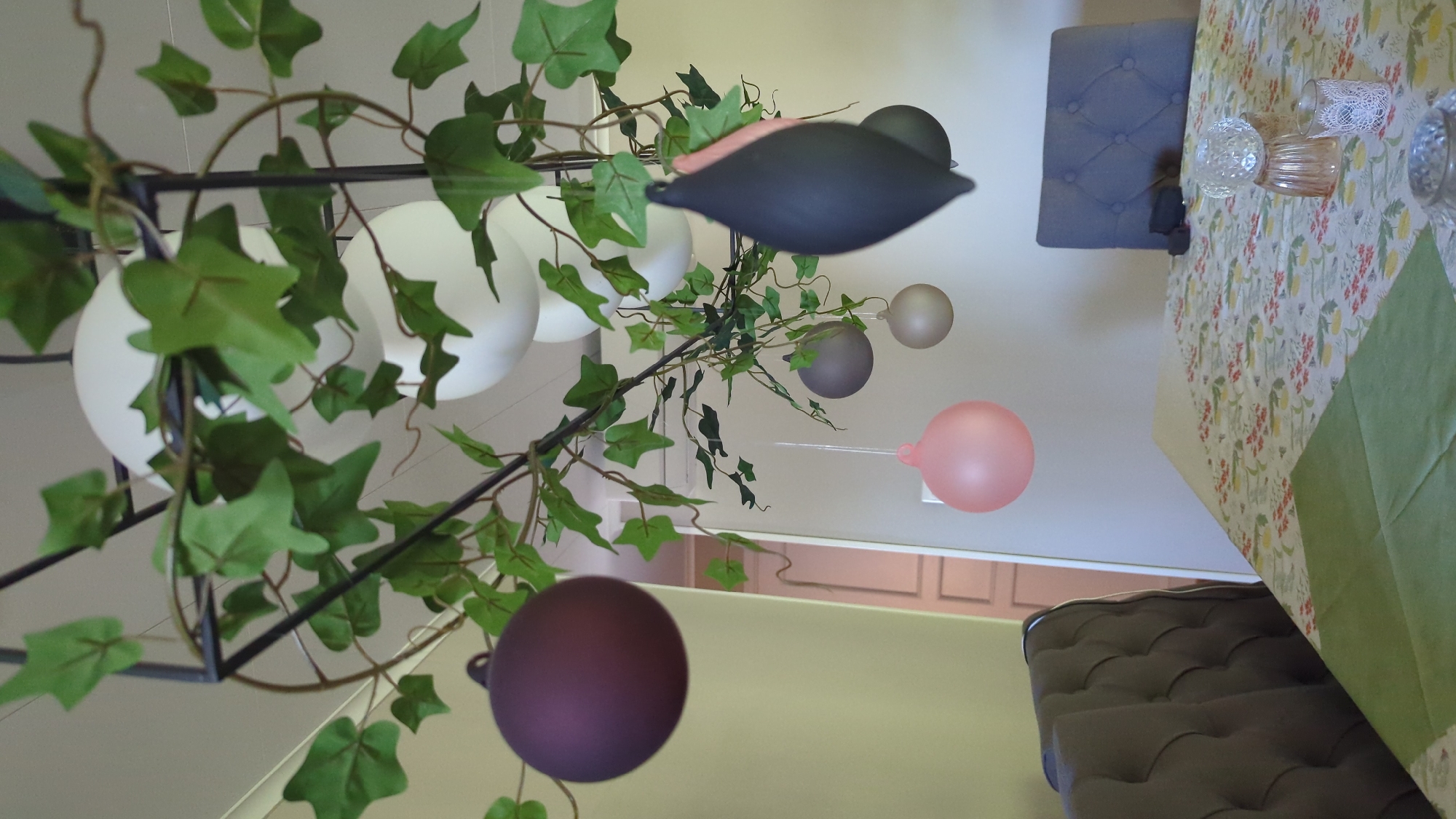 The image depicts an indoor setting, likely a dining room, adorned with a decorative hanging arrangement. The centerpiece of this arrangement appears to be a geometric metal structure intertwined with artificial green ivy. Suspended from this structure are several decorative spheres in various colors, including deep purple, pink, grey, and light gold, as well as a dark, elongated teardrop-shaped ornament. Two large, white spherical light fixtures are also visible within the arrangement.
The scene includes a table covered with a patterned tablecloth featuring floral and fruit motifs. On the table, there are decorative glass items, possibly candle holders or vases, and a green fabric napkin. Two chairs are partially visible; one is a dark upholstered tufted chair on the left, and the other is a grey upholstered chair with a tufted back on the right. The background reveals a wall with a door and what appears to be an air conditioning unit.
The overall ambiance suggests a festive or decorative occasion, with the hanging ornaments and lighting contributing to the aesthetic. The location is identified as Kløfta, Norway, suggesting this is an interior space within that geographical area. There are no people visible in the image, and no actions are depicted.