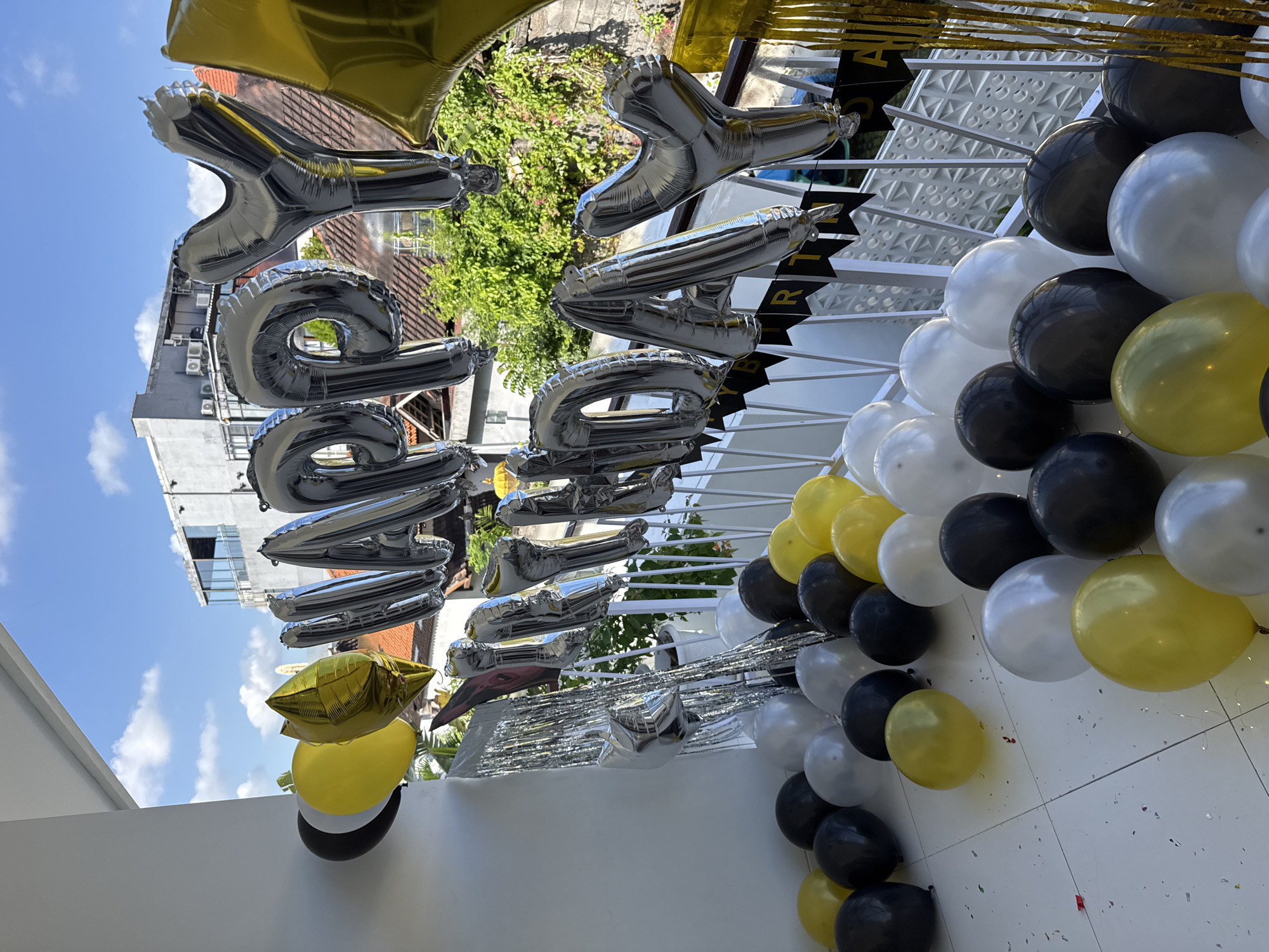 This image captures a balcony decorated for a birthday celebration in North Kuta, Indonesia. The focal point is a large silver balloon arch spelling out "HAPPY BIRTHDAY." Silver, gold, and yellow balloons are scattered across the balcony floor, some arranged in clusters, others lying individually. Behind the balloons, a white railing with a black banner displaying "HAPPY BIRTHDAY" is visible. The background shows a sunny day with blue skies and scattered clouds, overlooking rooftops and lush green trees. The balcony itself appears to be part of a residential or commercial building, with white walls and tiled flooring. No people are present in the scene, and no actions or interactions are depicted.