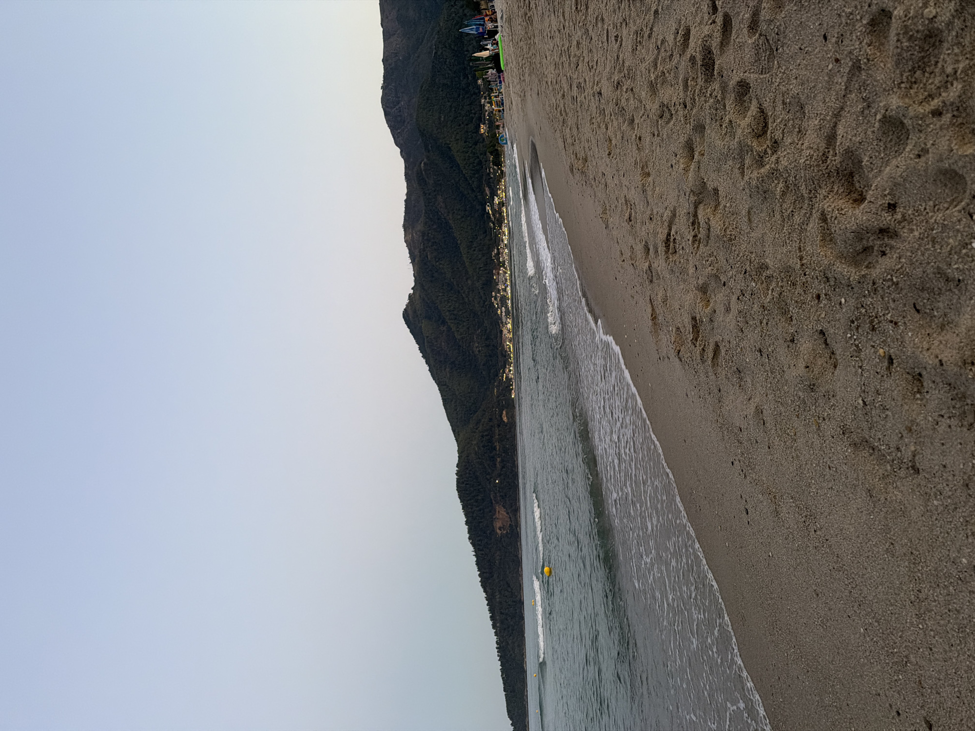 A tranquil beach scene unfolds in Golden Beach, Greece, bathed in the soft, muted light of dusk. The foreground is dominated by a sandy shore, marked with the imprints of numerous footprints, suggesting recent activity. Gentle waves lap the edge of the sand, their foam creating a delicate, white line against the tan grains.
Beyond the shoreline, the Aegean Sea stretches out, its surface rippling with subtle currents. Several yellow buoys are visible in the water, likely marking a swimming area or a navigational point. In the mid-ground, a lush green mountain range rises, its slopes densely covered with trees.
Along the base of the mountain, a cluster of buildings forms a small coastal town. Lights twinkle from the windows and streetlamps, casting a warm glow against the darkening landscape. The sky above is a gradient of pale blue and grey, transitioning from a lighter hue near the horizon to a deeper tone overhead, indicating the twilight hour. The overall atmosphere is one of peaceful serenity and natural beauty.
