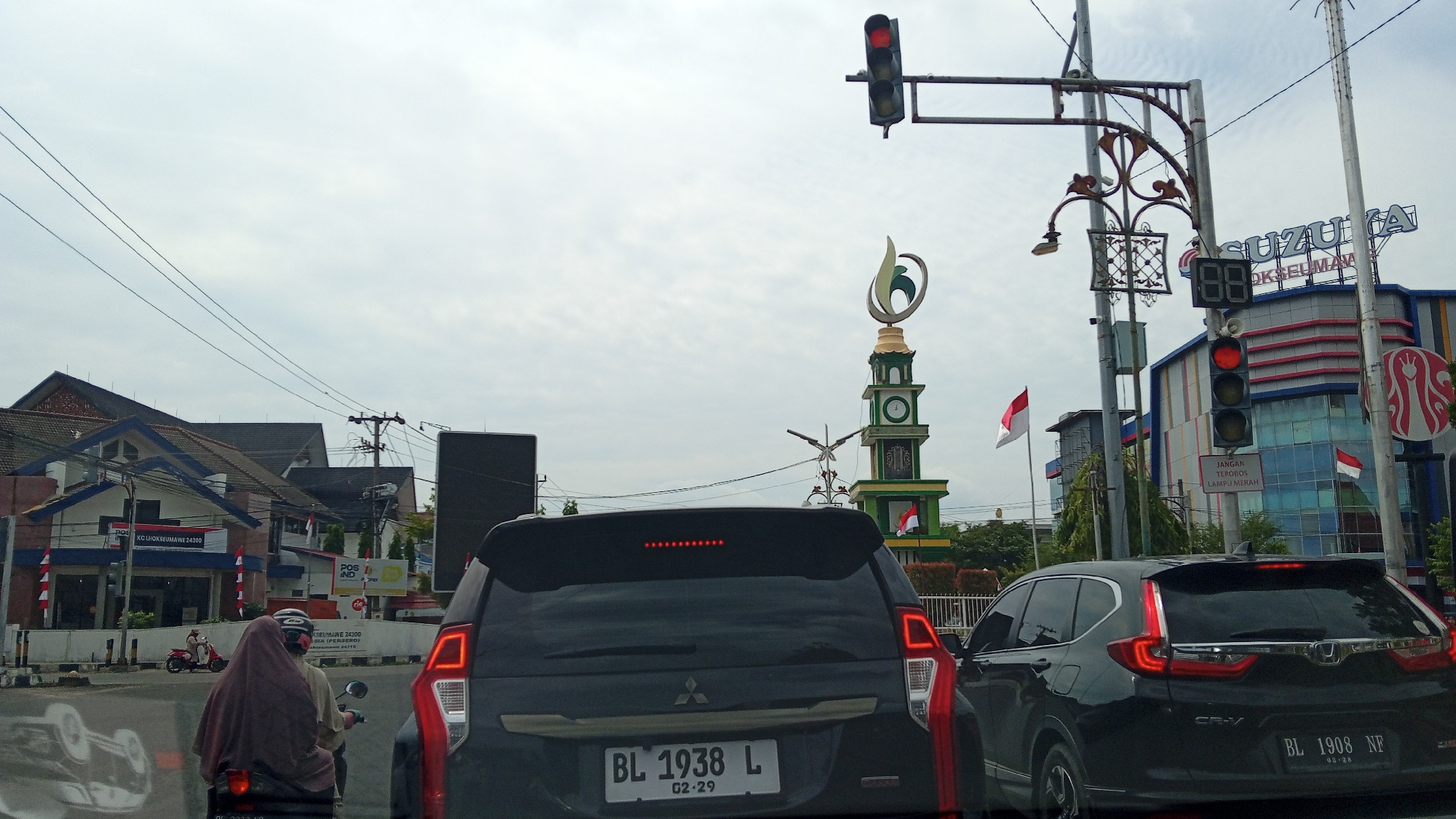 This is a horizontal, eye-level, medium shot captured from the interior of a vehicle. The scene depicts a traffic intersection in Lhokseumawe, Indonesia, under an overcast sky. In the foreground, the rear of a black Mitsubishi SUV with license plate BL 1938 L is prominently visible. To its right, the rear of a black Honda CRV with license plate BL 1908 NF is also visible. A motorcycle with two occupants, a driver wearing a helmet and a passenger in a pink hijab and brown headscarf, is positioned to the left of the SUV.
The intersection features a traffic light with a red signal illuminated and a digital countdown timer displaying "88." A decorative clock tower, adorned with green and white elements and a stylized flame-like emblem at its apex, stands in the middle of the intersection. Indonesian flags are displayed on poles near the clock tower.
Buildings line the sides of the street. On the left, a building with a dark tiled roof and white and blue accents is partially visible. A banner with "POS IND" and "KC Lhokseumawe 24300" suggests it is a post office. On the right, a large, modern building with a blue, red, and white facade is labeled "SUZUKI." Additional signage is present, including a red sign with white text that reads "JANGAN TEROBOS LAMPU MERAH" (Do not run the red light). Power lines crisscross the sky. The overall impression is of a busy urban intersection during daylight hours.