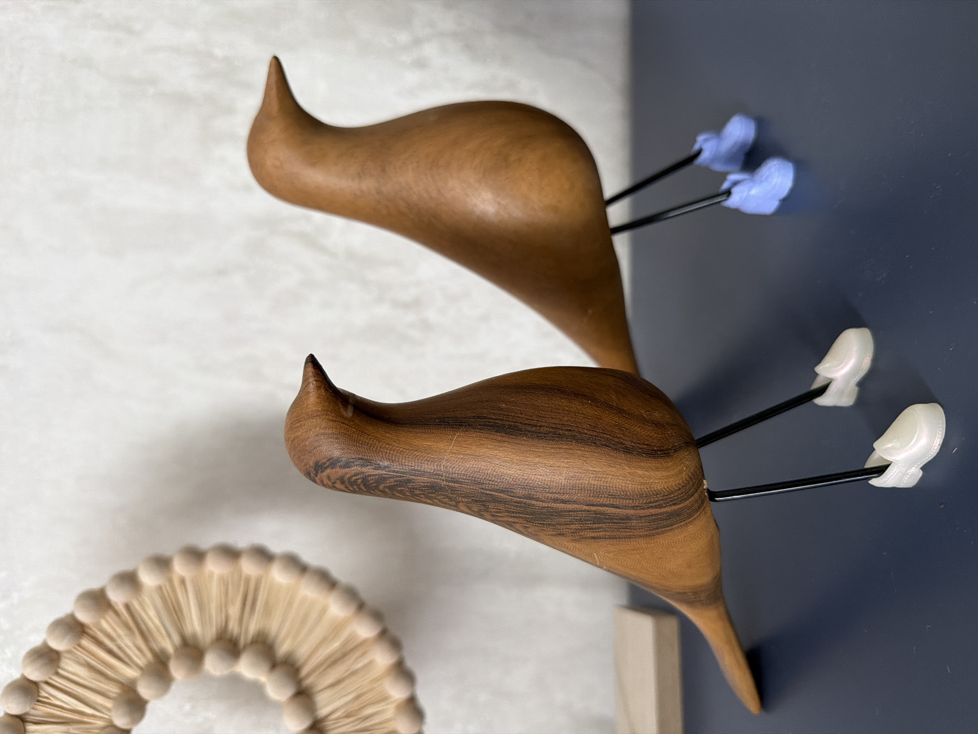 Two wooden bird figurines are displayed on a dark blue surface. The bird on the left is closer to the camera, facing left with its head slightly turned towards the right. It has a visible wood grain pattern. The bird on the right is further back, facing right. Both birds have thin black legs and wear miniature shoes on their feet. The bird on the left wears iridescent white shoes, while the bird on the right wears light blue shoes. In the background, a textured white wall is visible, with a circular, fan-like wooden decoration on the upper left side of the image. A light-colored rectangular object is partially visible on the left side of the surface. The scene is indoors. There is no information in the image to determine its location in Novosaratovka, Russia.