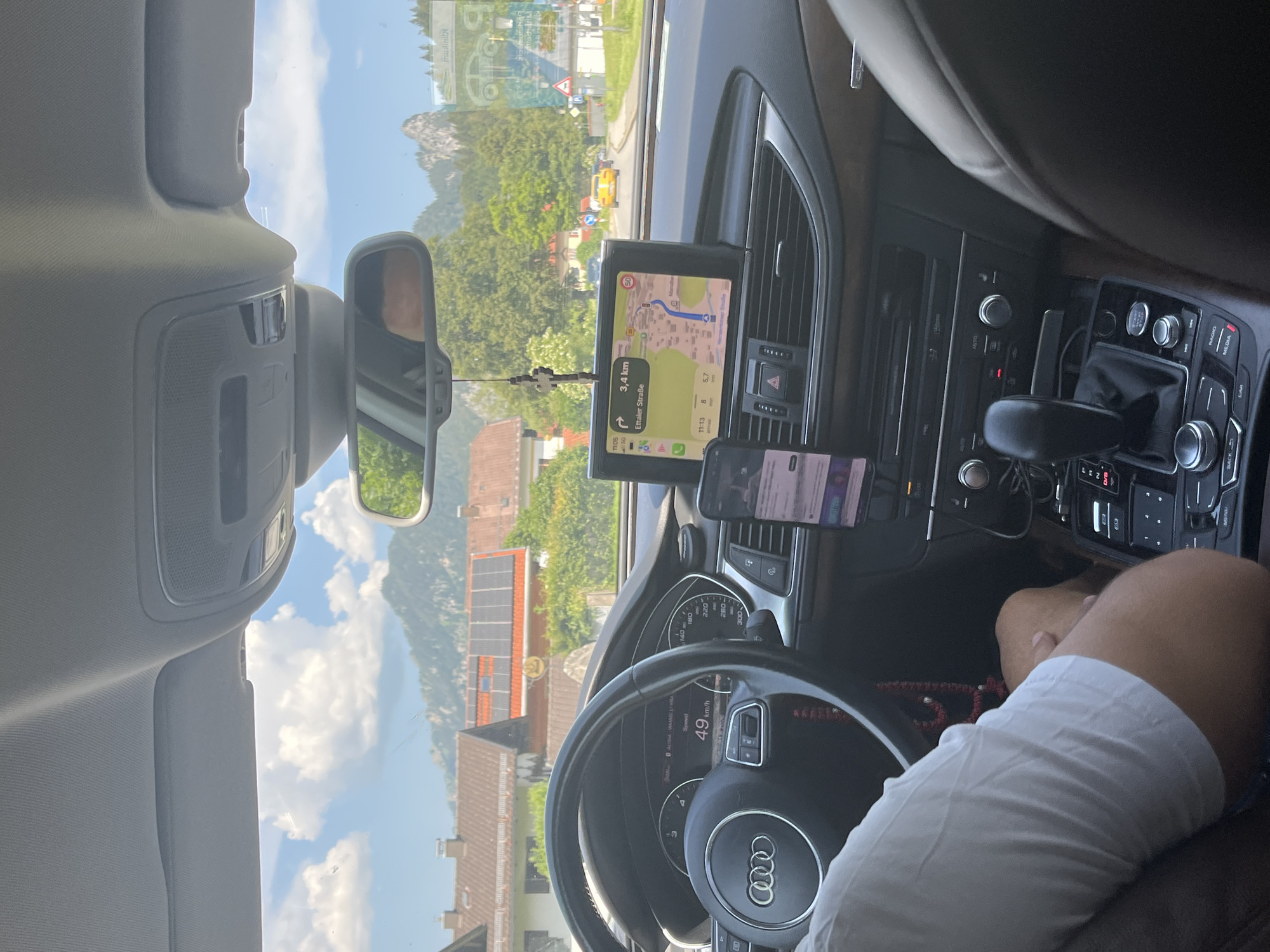 The image is taken from the interior of a car driving through the town of Oberau, Germany. The driver, a person with fair skin wearing a white t-shirt, is partially visible on the left, holding the steering wheel. The car's dashboard features a navigation screen displaying a map and route information, indicating a destination of "Ettaler Straße" and a distance of 3.4 km. The car's speed is shown as 49 km/h. Outside the car, typical Bavarian architecture with red-tiled roofs and solar panels is visible. In the background, lush green trees and mountains under a partly cloudy sky suggest a scenic, possibly rural or suburban, setting within Oberau. A yellow car is visible driving ahead on the road.