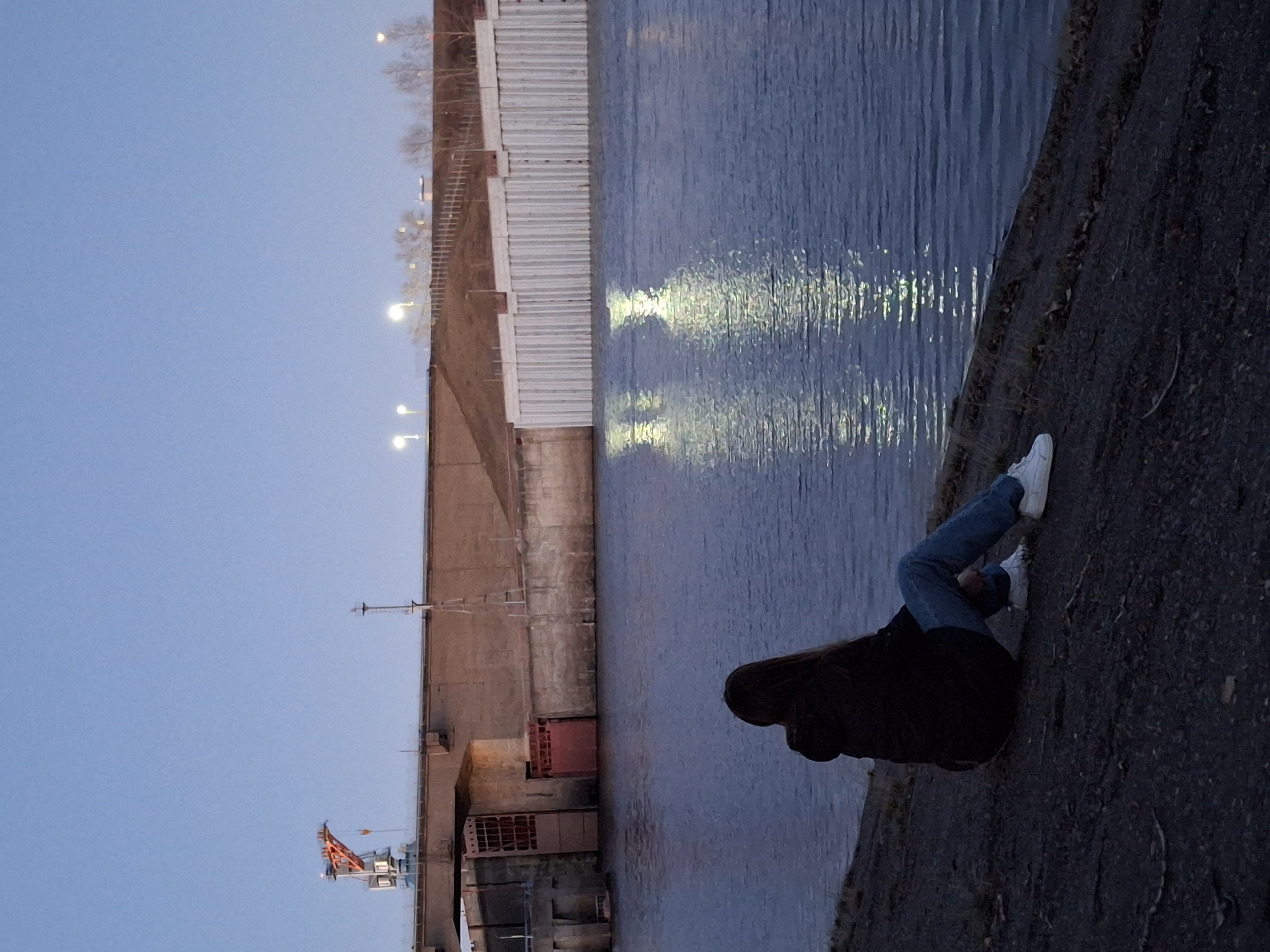 A woman sits on a concrete bank near a body of water, possibly the Kama River, in what could be Chaikovsky, Russia, based on architectural clues such as the concrete structure lining the opposite bank and the style of street lighting. The woman is wearing jeans, white sneakers, and a dark jacket with her hood up. She is facing the water, her posture suggesting contemplation. The lighting indicates either dusk or dawn. On the opposite bank are industrial-looking structures, possibly related to river transport or energy production, including a gantry crane. Streetlights line the far bank. The water's surface reflects light, suggesting the presence of lights. The overall scene has a somber, quiet atmosphere.