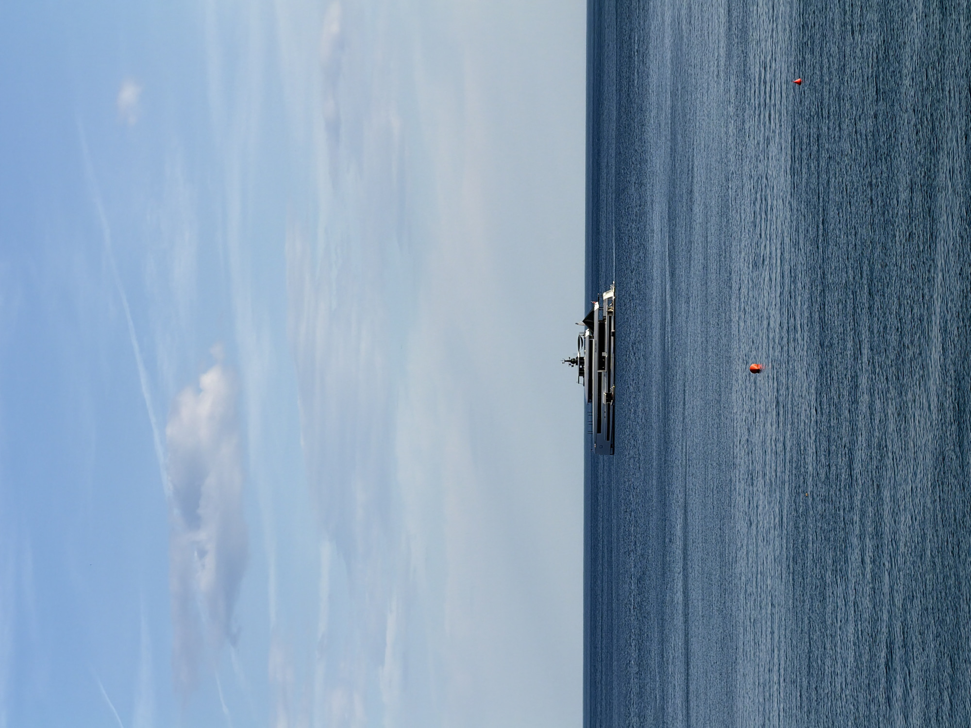 A vertical wide shot depicts a partly cloudy sky, ocean, and a luxury yacht, at an undetermined location. The left side of the image shows a light blue sky with wispy clouds. The right side shows dark blue ocean water. A large yacht is centered in the image, between the sky and the ocean. Two red buoys are visible in the water.