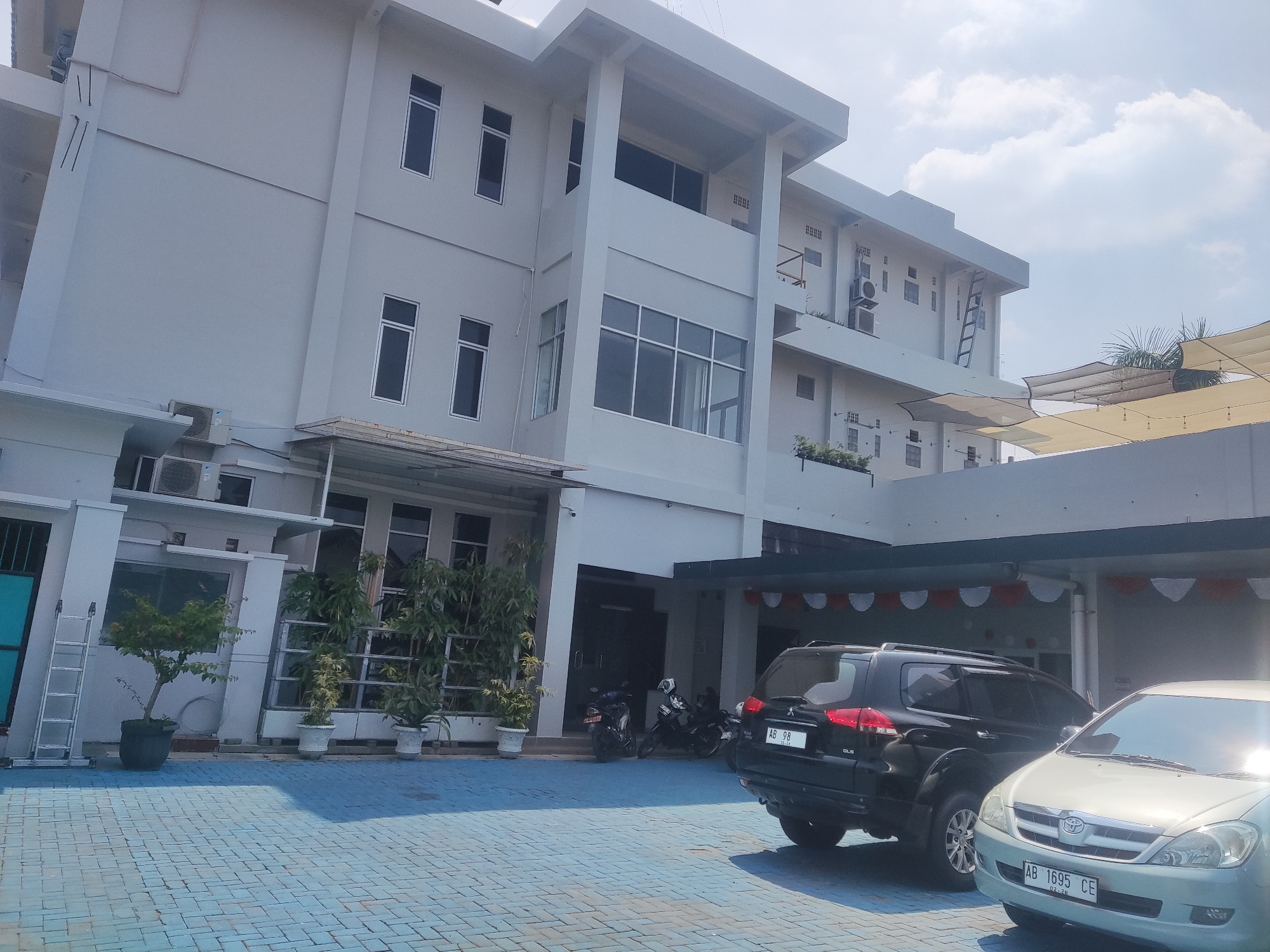 The image shows a light gray, multi-story building with a modern architectural design, likely an office building or a hotel, situated in Trihanggo, Indonesia. The building features numerous windows with dark frames, several air conditioning units mounted on the exterior walls, and a covered walkway with decorative banners. In front of the building, a blue tiled courtyard is visible, with potted plants and a silver ladder leaning against the wall. Two cars, a black SUV and a silver minivan, are parked in the courtyard. In the background, several motorcycles are parked under a covered area. The sky is partly cloudy, indicating daytime. The scene appears to be a well-maintained and organized exterior area of a commercial or institutional property.