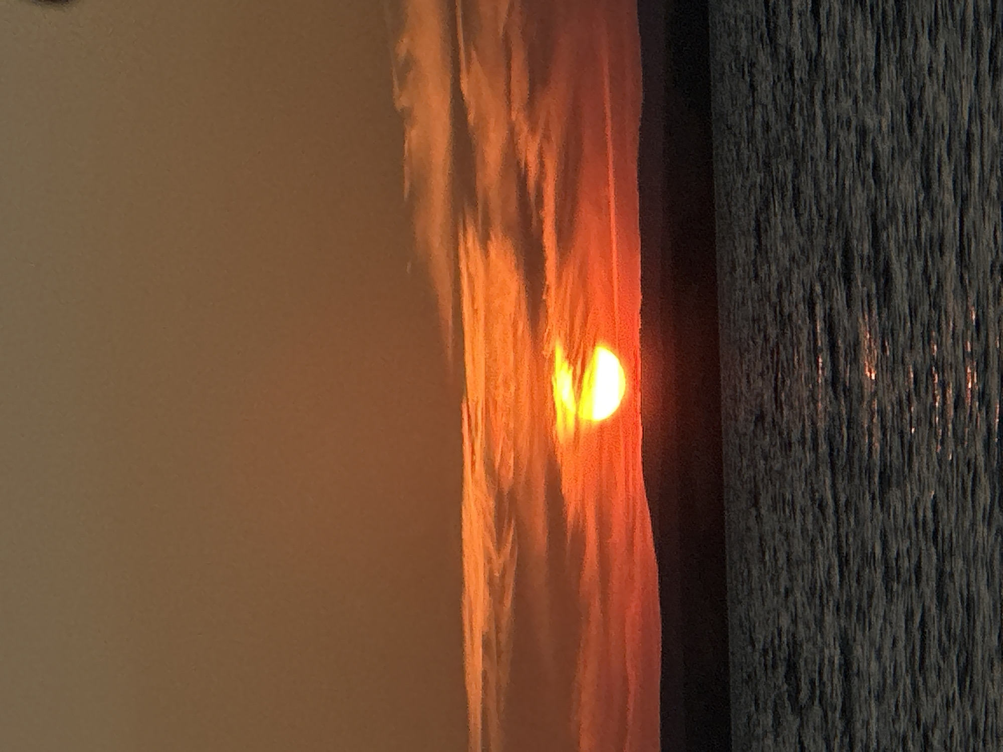 The image depicts a serene sunset over a body of water, likely the dam of the Zeysky Reservoir near Energetik, Russia. The sun, a brilliant orb of orange and yellow, is partially obscured by the horizon, casting a warm glow across the sky and illuminating the wispy clouds. The water's surface is calm, reflecting the vibrant hues of the sunset, with gentle ripples suggesting a light breeze. A dark, silhouetted landscape forms the distant horizon, separating the sky from the water. No people or artificial structures are visible, emphasizing the natural beauty of the scene. The overall impression is one of tranquility and natural splendor.