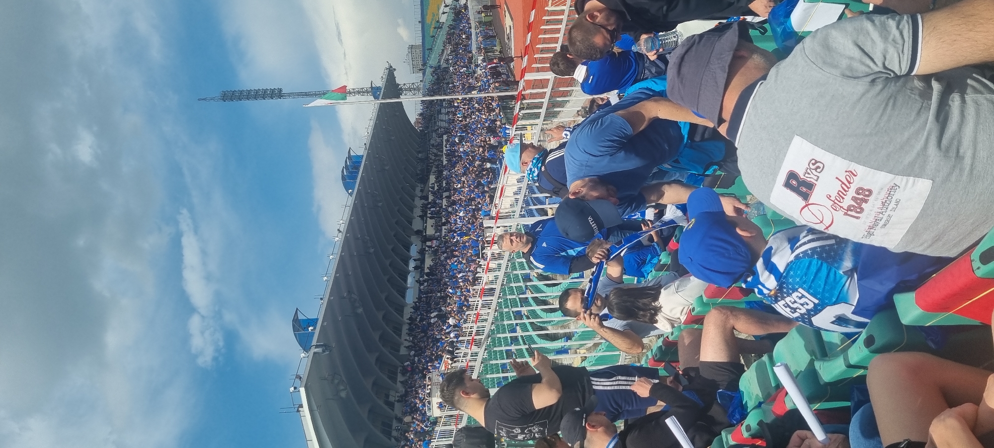 A vertical medium shot shows a stadium and the people sitting in the green seats. The stadium is on the left side. The sky is blue and white and in the upper left side. The top of the stadium has a flag on a tall pole. The colors on the flag are red, white, and green. The people sitting are all ages, wearing casual clothing, and facing the stadium. In the lower right corner is a man wearing a grey shirt with a white and red logo. The city is unknown.