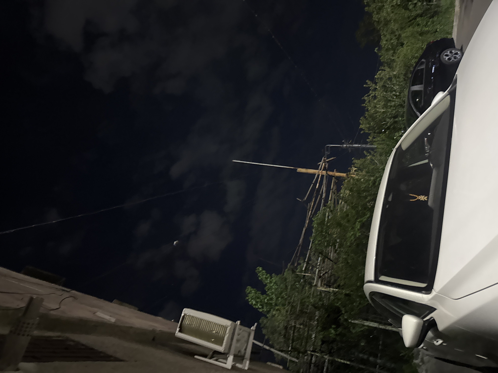 The image captures a night scene in Yerevan, Armenia. A white car dominates the right side of the frame, partially obscuring a black car parked behind it. To the right of the cars is dense greenery suggesting trees. In the center, bamboo scaffolding is visible along with a light fixture on a building. The sky is dark and partly cloudy. The image is taken from a low angle.