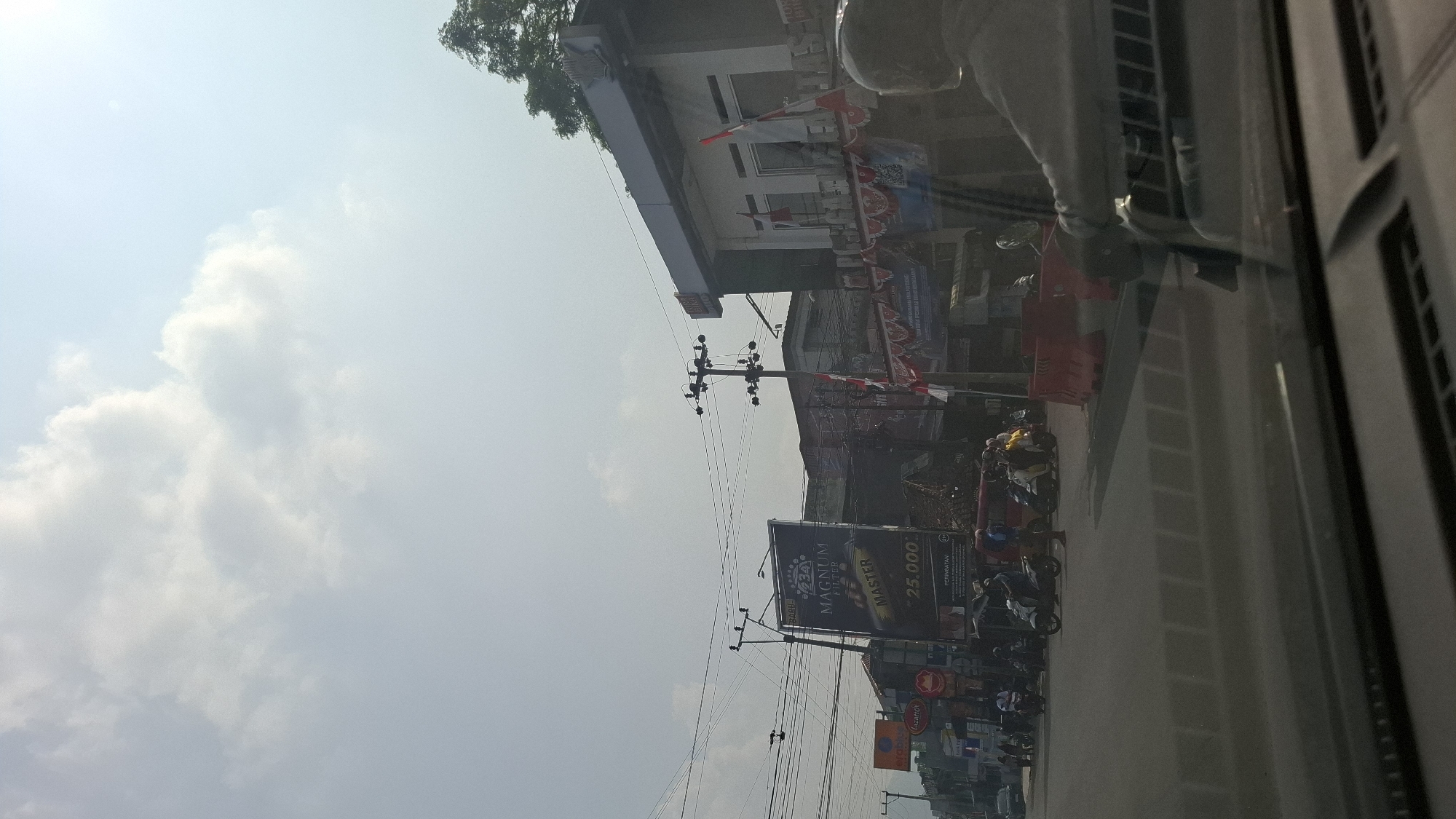 The image captures a street scene in Cikaroya, Indonesia, under a bright, partially cloudy sky. In the foreground, the dashboard of a vehicle is visible, indicating the photograph was taken from inside a car. The road ahead is populated with several motorcycles and a van. To the left, a large billboard advertises "MAGNUM FILTER MASTER" with a price of "25.000". Further down the street, various buildings line the road, adorned with Indonesian flags and banners. One building in the mid-ground has a prominent sign and windows decorated with flags. Electrical wires crisscross the sky, supported by utility poles. To the right, a person, likely a passenger in the vehicle from which the photo is taken, is partially visible, facing away from the camera. Orange traffic barriers are set up on the right side of the road. The overall impression is that of a bustling street on a sunny day in an Indonesian city.