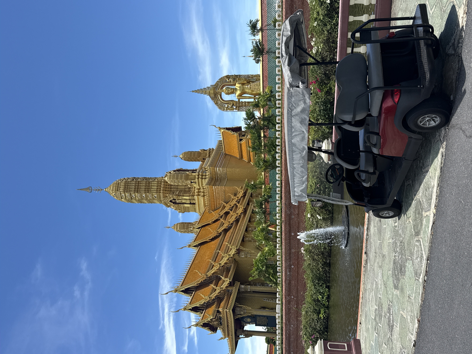 The image shows Wat Asokaram, a Buddhist temple in Bang Pu Subdistrict Municipality, Mueang Samut Prakan District, Samut Prakan, Thailand, with a golf cart in the foreground. The temple's golden spires and intricate architecture are visible against a blue sky with wispy clouds. A golf cart sits to the right of the frame, with a cover partially obscuring its details. A small pond with a fountain separates the temple complex from the golf cart. The ground is paved with stone tiles, and a grey asphalt road runs along the right edge of the picture.