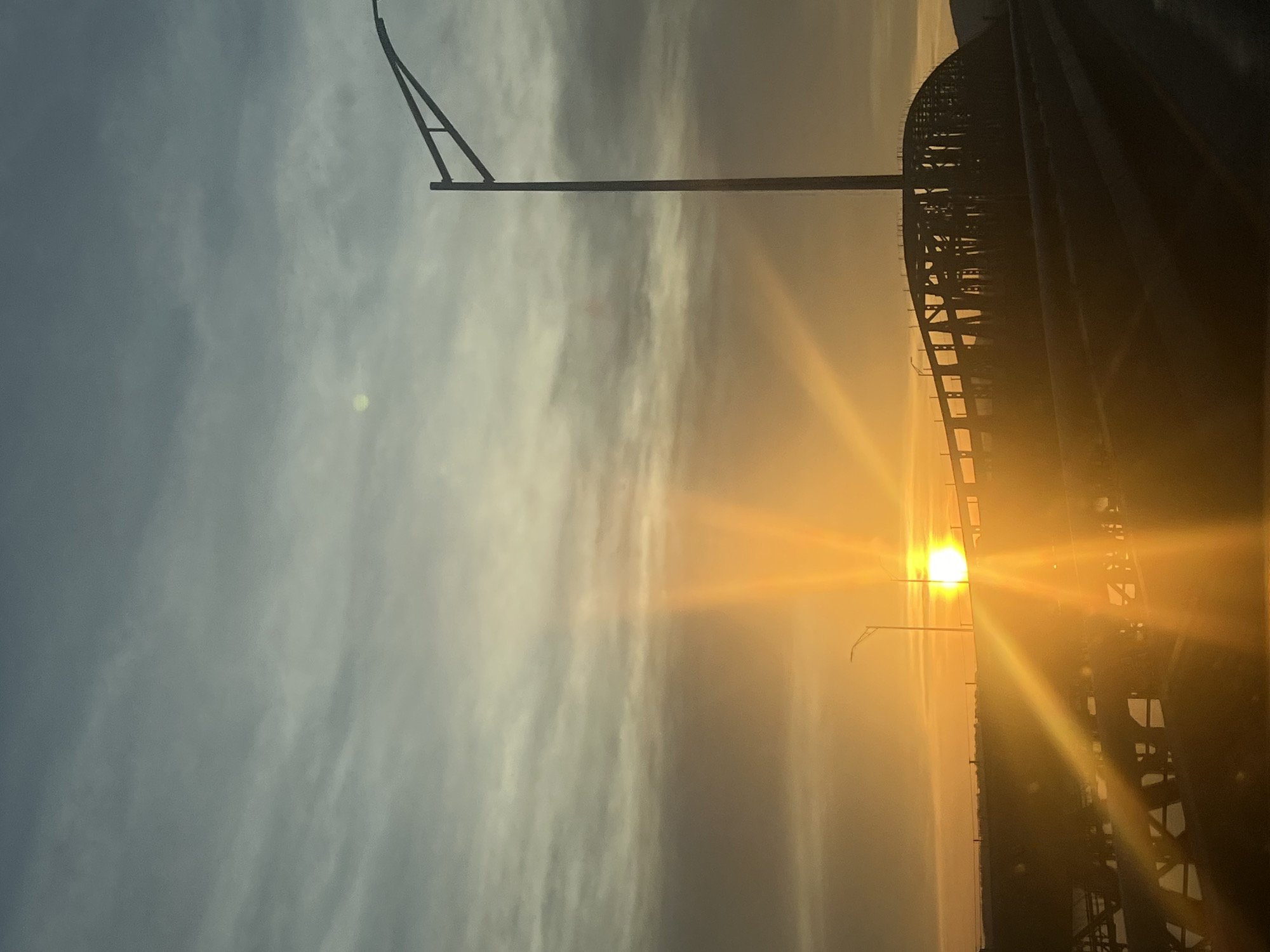 Here is a description of the image:
The image depicts a sunset view from the City of Newburgh, United States of America, with the sun prominently positioned behind a bridge structure. Silhouetted against the bright sunlight, the bridge appears to be made of steel with a complex grid pattern. To the left of the image, a streetlight pole is visible. The sky displays a gradient of colors, shifting from a blue-gray hue near the upper left corner to an orange and yellow glow near the horizon where the sun is located. The sunlight reflects and shines through the bridge supports.