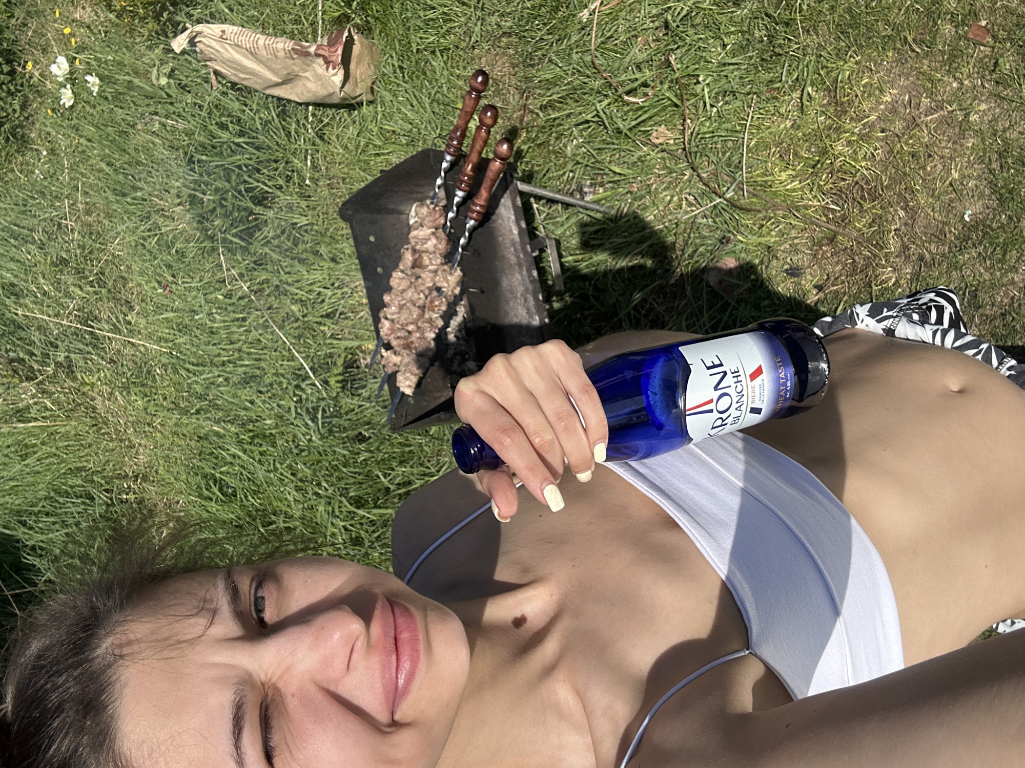A person wearing a white bikini top lies on the grass, holding a blue Kronenbourg 1664 Blanc beer bottle. A barbecue grill with skewers is nearby. A bag is also visible on the grass. The location is assumed to be outdoors, possibly in Khokhryaki, Russia.