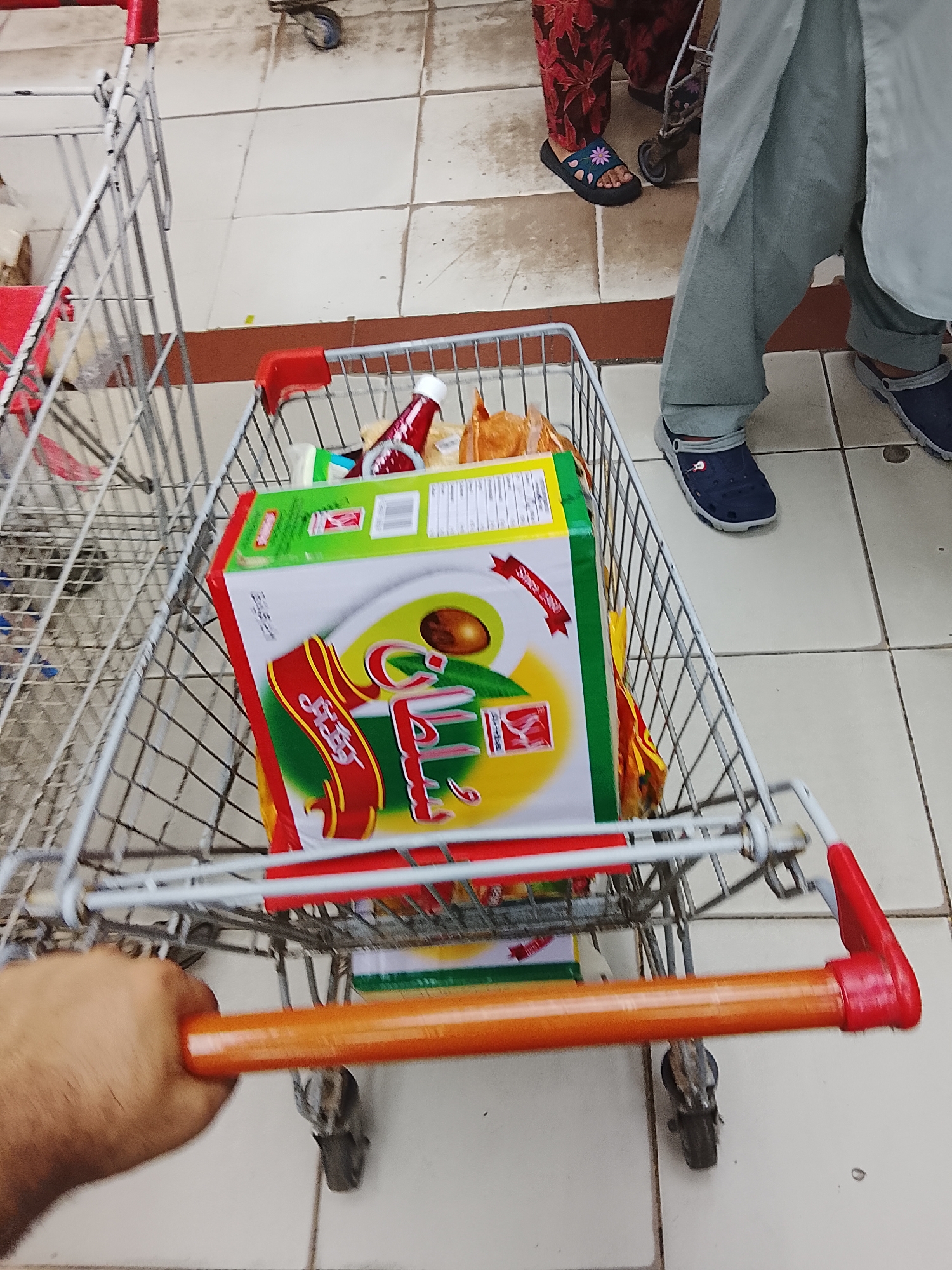 In a supermarket located in Gulgasht Colony, Pakistan, a first-person view shows a hand pushing a shopping cart filled with grocery items. The cart contains a large box, a bottle of ketchup, and other packaged goods. Two individuals are visible in the background, one wearing a floral-patterned dress and sandals, the other in a grey shalwar kameez and blue slip-on shoes. The supermarket floor is tiled.