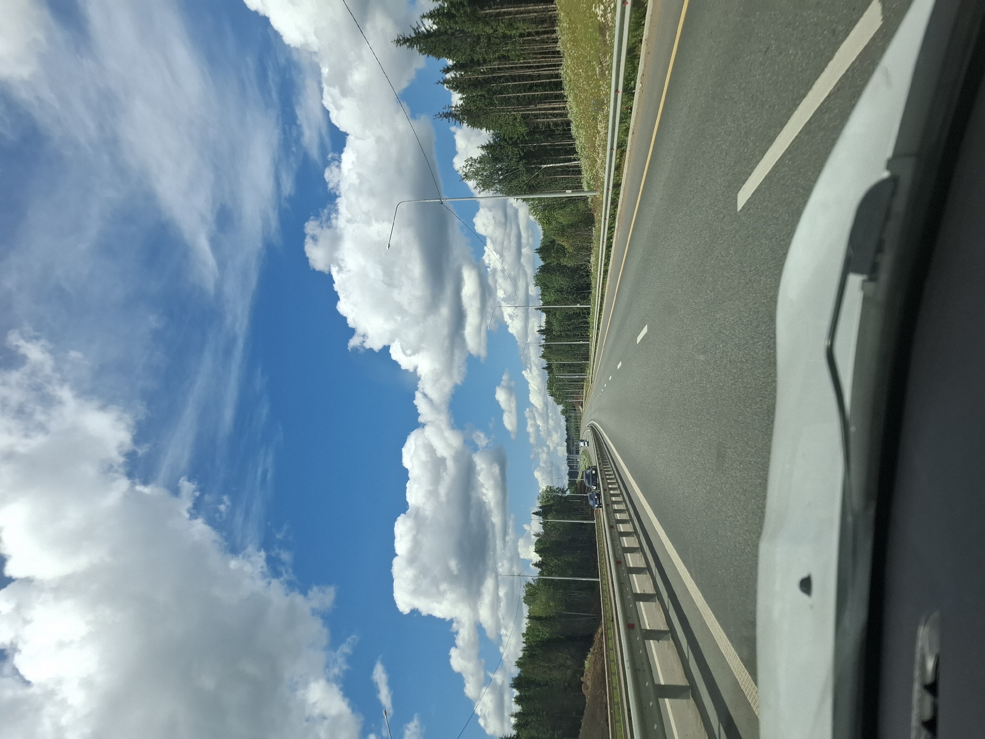 This is a POV shot from a vehicle driving on a highway. The highway has two lanes in each direction, separated by a median. Guardrails line the side of the road, and trees and a grassy embankment are visible beyond the guardrails. The sky is bright blue with scattered white clouds. There are streetlights lining the highway. A few cars are visible on the road ahead. The foreground shows the dashboard and windshield of the vehicle. The image was taken in Urazgil'dy, Russia.