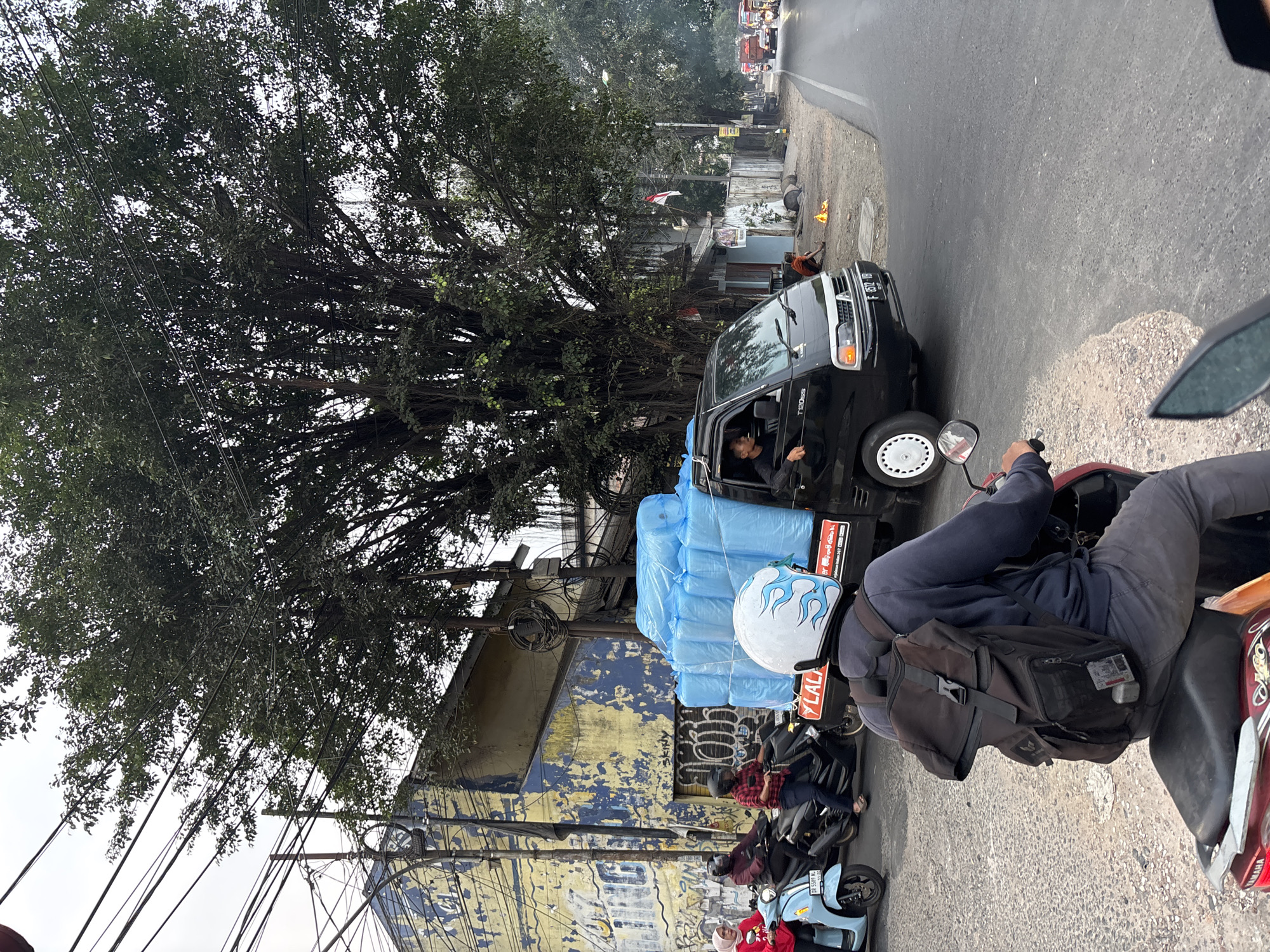The image shows a street scene in Bekasi, Indonesia, with a focus on a black pickup truck carrying a large load of blue-wrapped items, possibly mattresses or similar goods, secured to its bed. The truck is positioned on the right side of the frame, in the middle of the road, and its driver is visible through the open driver's side window. Several motorcycles are also present. In the foreground, a person wearing a backpack and a helmet with a flame design rides a motorcycle, their back to the viewer. To the left of the truck, other motorcycles are stopped or moving. One light blue scooter has two passengers, a woman in a red headscarf and a person in a dark jacket. A black motorcycle with a rider in a plaid shirt is also visible. Behind the vehicles, a dilapidated building with peeling yellow and blue paint stands prominently, adorned with graffiti. Large trees with dense foliage, characteristic of tropical urban environments, dominate the upper portion of the image, with numerous electrical wires crisscrossing overhead. The overall atmosphere suggests a bustling street in a city with a mix of older infrastructure and active daily life.
