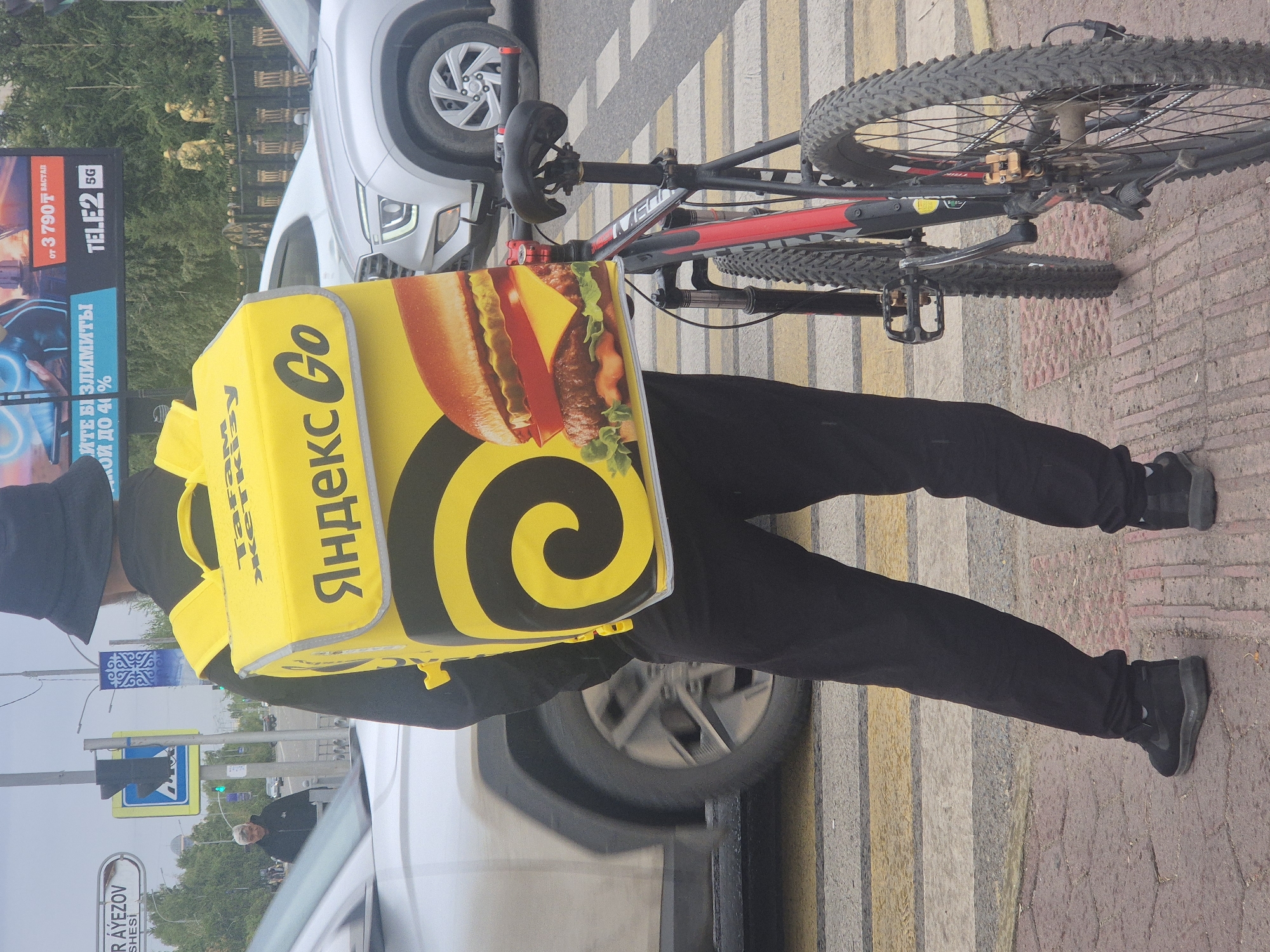 In Kokshetau, Kazakhstan, a person wearing a black bucket hat and black clothing stands on a pedestrian crossing. They are carrying a large yellow insulated backpack that advertises "Yandex Go" and features an image of a hamburger. A bicycle with a red frame and black tires is parked next to the person, leaning against the backpack. The background shows a street with moving vehicles, including a silver SUV, and traffic signals. A billboard advertising "TELE2 5G" is visible in the distance, along with some decorative fencing and statues.