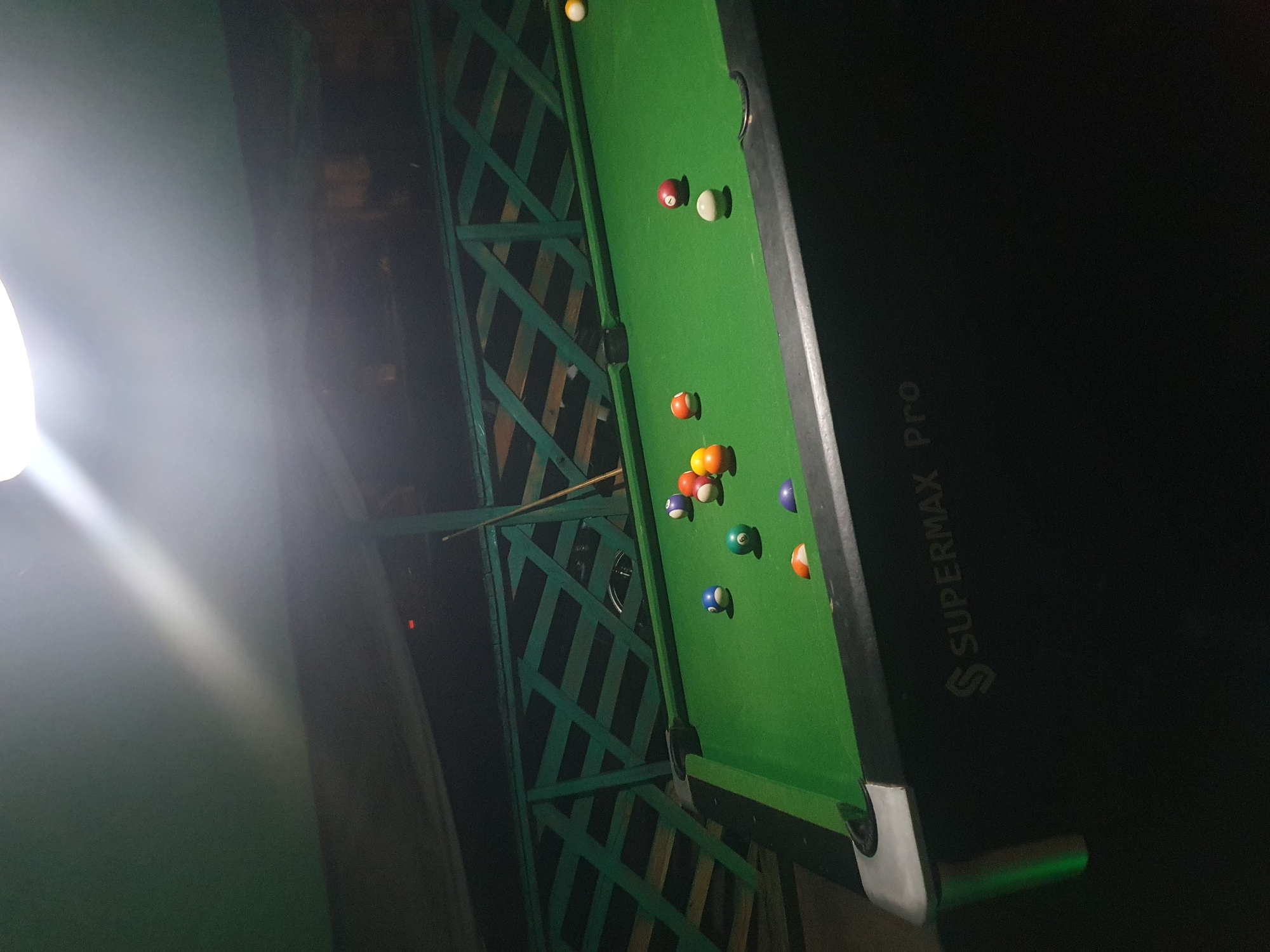This is an exterior shot of a Supermax Pro brand pool table, set up outdoors under a covered, open-air structure. The pool table is green, with several billiard balls scattered across its surface, including a white cue ball, solid colored balls numbered 1 through 8, and striped balls. The structure has a green wooden lattice railing in the background. The overall scene is dimly lit, with a bright light source illuminating the upper left portion of the frame. It is difficult to determine the specific location within Lokoja, Nigeria, from the visual evidence provided.