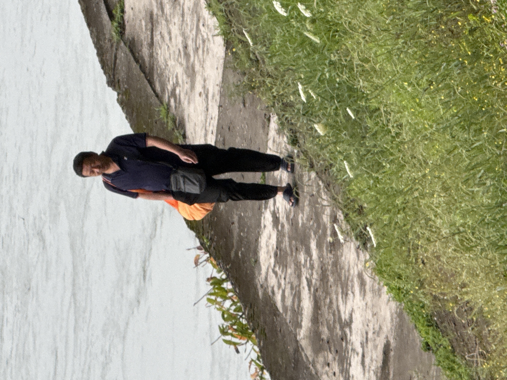 A medium shot depicts a man wearing a black t-shirt, black pants, and sandals. He stands on a stone pathway with a dark bag slung across his shoulder. The pathway is next to a body of water and extends towards a grassy area. The scene is located in Jiangjin, China.