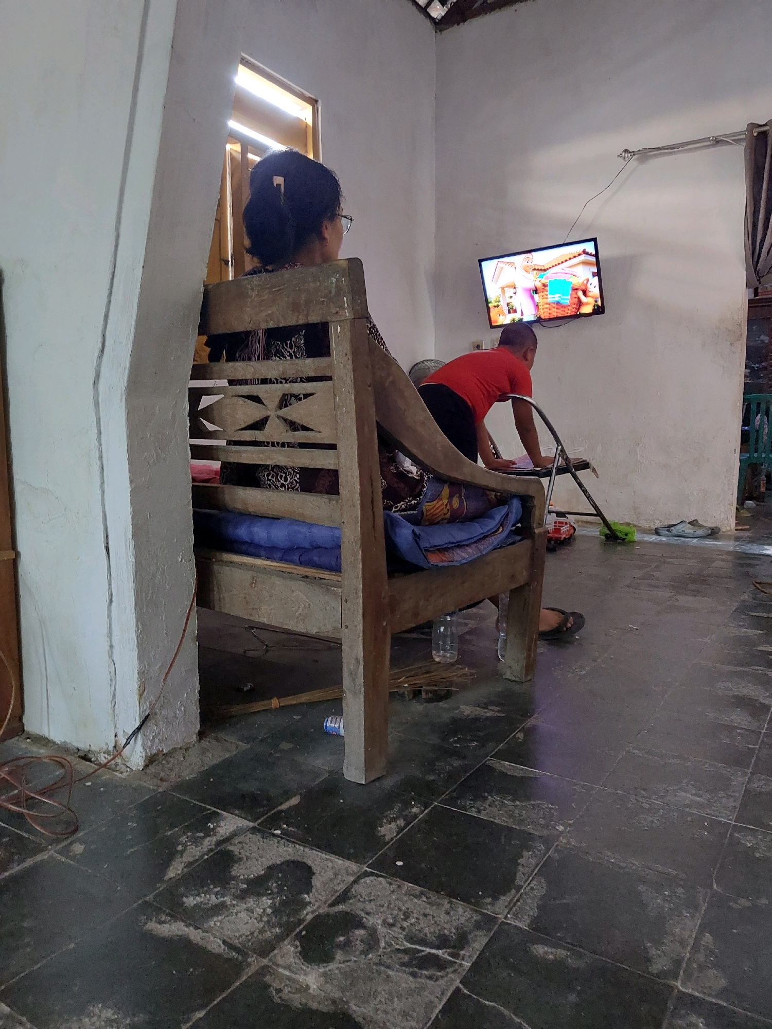 In a room with a light-colored wall and dark tiled floor, a person sits on a wooden chair facing away from the camera. The person has dark hair pulled back and wears glasses. In front of the chair, a child in a red shirt and dark pants bends over a collection of toys, including a red toy truck and a green toy. A television on the wall displays a colorful animated scene. Various items are scattered on the floor, including a broom, a bottle, and footwear. The room appears to be a domestic setting, likely in the city of Sarangan, Indonesia.