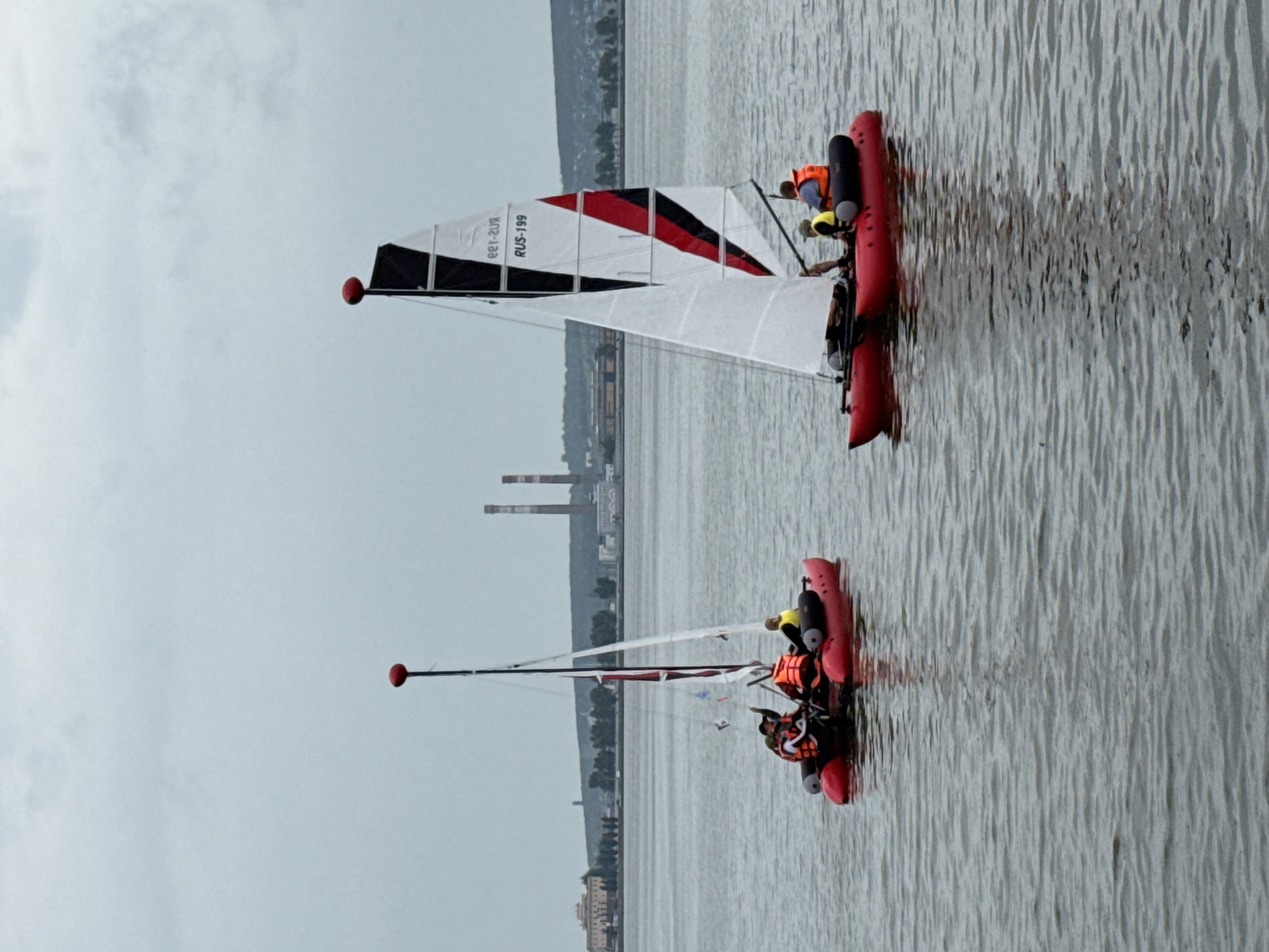 Two red inflatable catamarans with sails are on a body of water, likely the Votkinsk Reservoir, in Votkinsk, Russia, under an overcast sky. The catamaran on the right has a tall, triangular sail with black, white, and red sections, displaying the text "RUS-199" and some mirrored Cyrillic text. It carries two individuals, one wearing an orange life vest and the other a yellow and orange life vest. The catamaran on the left has a simpler sail and carries three individuals, all wearing orange life vests. In the background, a shoreline with trees and buildings, including industrial structures with tall chimneys, is visible. The water is calm, with ripples visible.