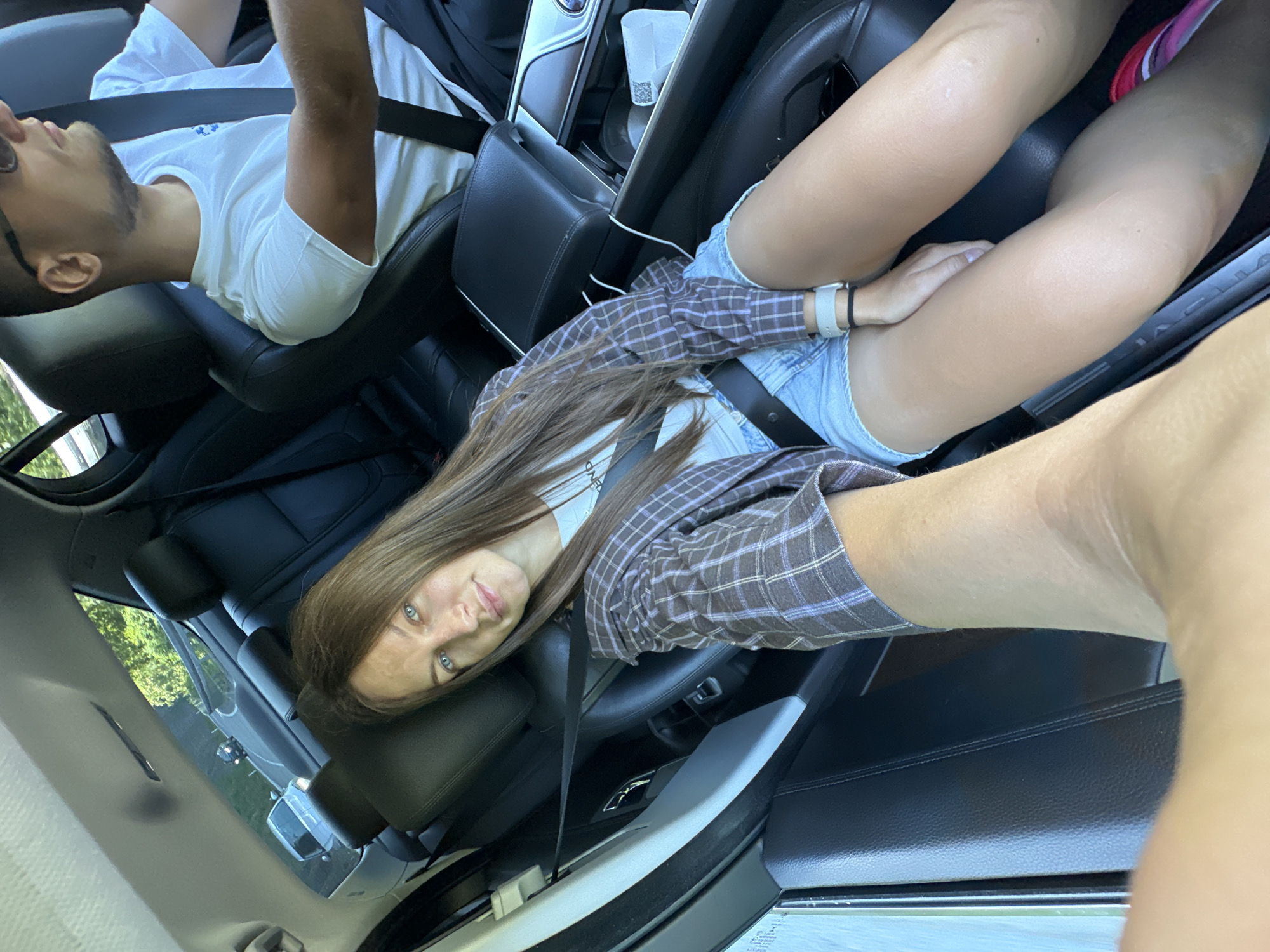The image shows three people in a car. In the foreground, a person's arm and shoulder are visible, obscuring part of the scene. The primary subject is a young woman with long brown hair, wearing a plaid shirt and denim shorts. She is looking at the camera with a neutral expression. She is sitting in the passenger seat of the car. Next to her, a man wearing a white t-shirt and sunglasses is seated in the driver's seat. His head is tilted back, and his eyes are closed, suggesting he may be asleep or resting. The interior of the car is dark, with black leather seats. The setting appears to be daytime, with sunlight filtering into the car, indicated by the shadows and the bright green foliage visible through the windows. The car is parked, possibly on a road or in a scenic location given the presence of trees. The overall mood is relaxed, possibly a road trip or a casual outing. Visible text includes "JAGUAR" on the car door sill. The image was taken in Jabuka, Montenegro.