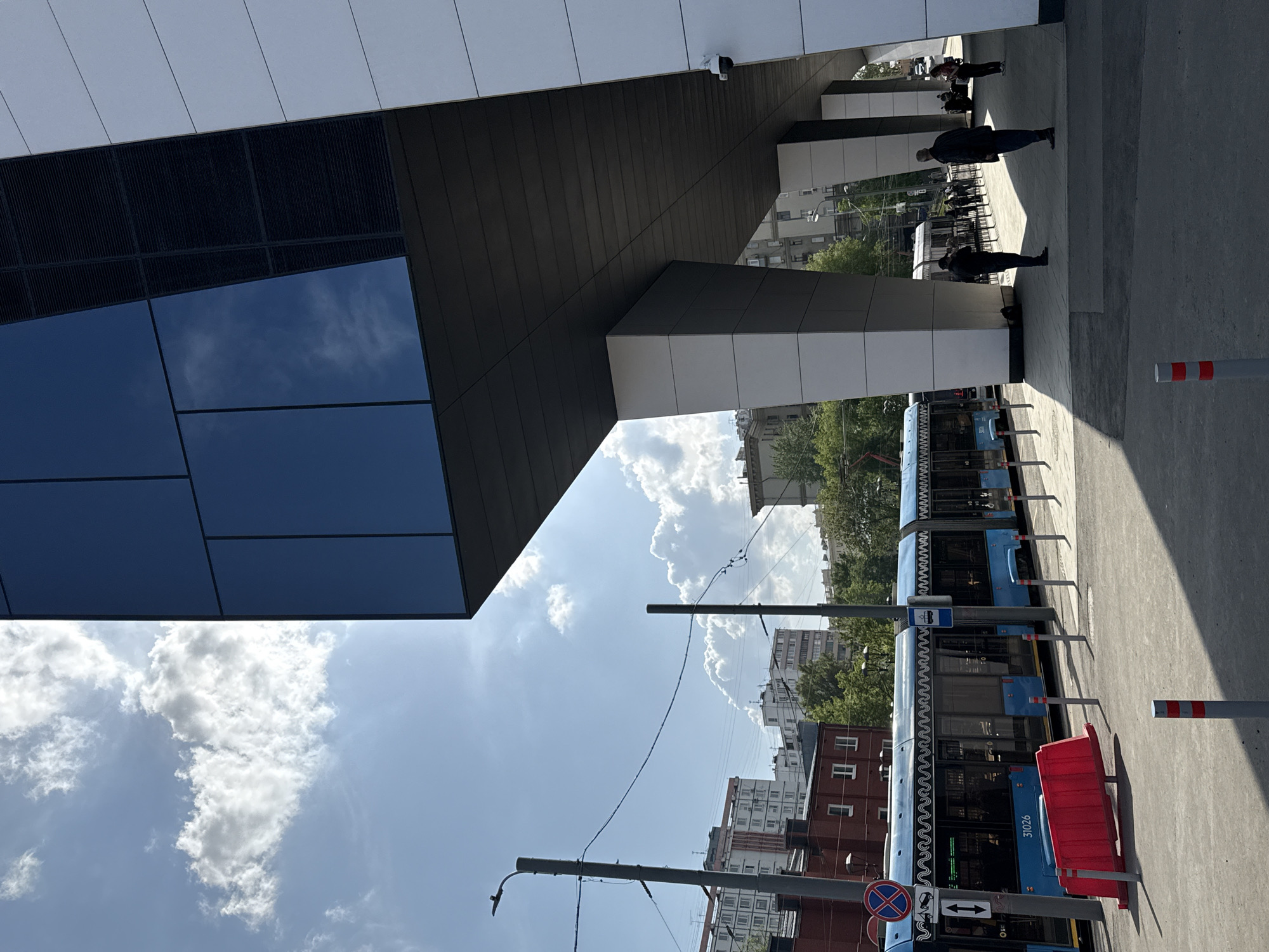 This is an outdoor shot taken in Moscow, Russia, showcasing the exterior of what appears to be a modern building. The architectural elements feature a dark-paneled section reflecting the blue sky with fluffy clouds, juxtaposed with segments of white and gray structures. The sky is visible both directly and in the building's reflections. Underneath the building's overhang, several individuals are walking and standing, indicating pedestrian traffic. A light blue tram is parked parallel to the walkway, complete with its identifying number, 31026, and a red protective barrier. Street furniture such as signposts and pedestrian markers are visible. The wider environment includes trees and apartment buildings, characteristic of an urban Russian setting.