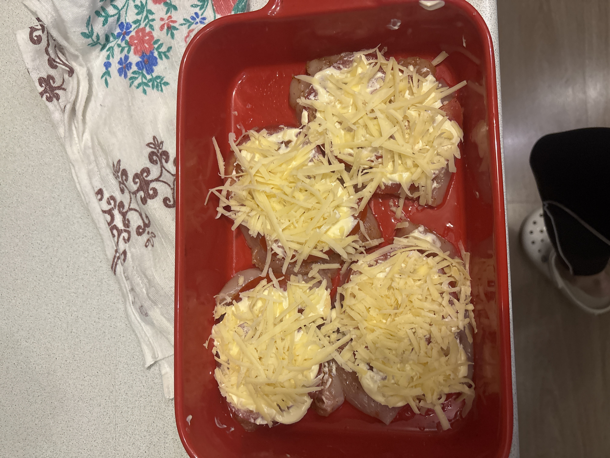 The image shows a red baking dish containing four pieces of meat topped with mayonnaise and shredded cheese. The dish is placed on a countertop, beside a white cloth with a floral pattern. A black garment and a shoe are visible in the background. The location is in the city of Chaikovsky, Russia.