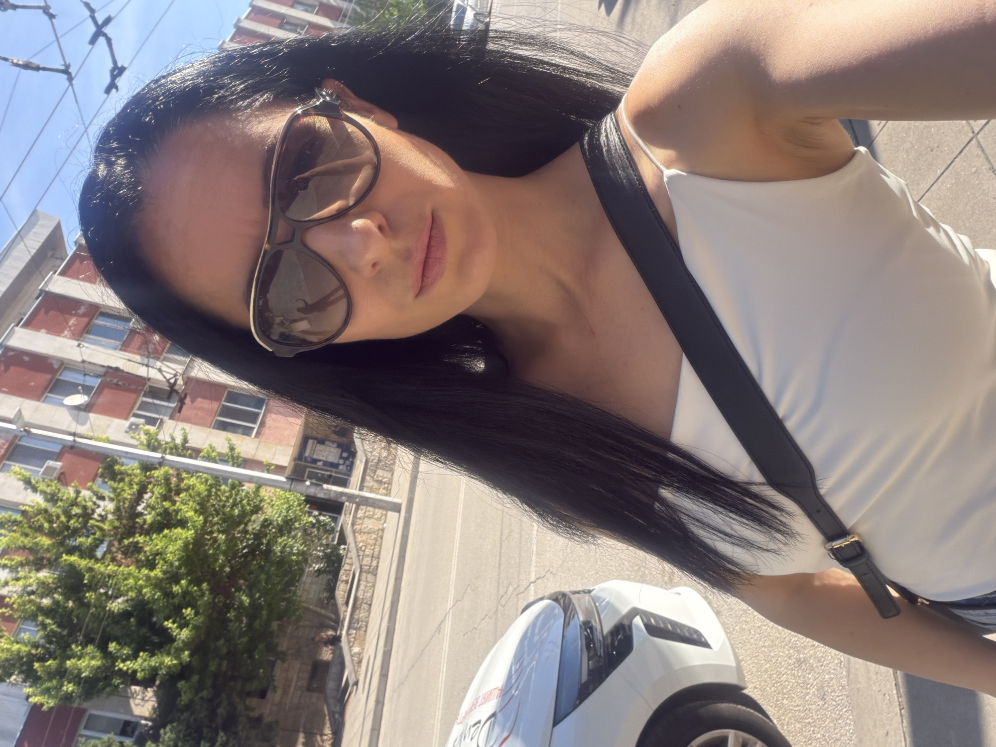 The image is a tilted outdoor selfie of a young woman in Pleven, Bulgaria, captured on a bright, sunny day. The woman, with long, straight dark hair, wears large brown-tinted sunglasses and a white camisole top with thin straps. A black shoulder strap with a gold buckle crosses her chest. She holds the camera at an angle, looking towards it with a neutral expression.
In the background, under a clear blue sky, are urban elements. Buildings with several windows and reddish-brown and gray facades are visible, along with lush green trees. Overhead lines, likely for public transport like a trolleybus or tram, can be seen against the sky. A portion of a white car with some red text, possibly "DRIVE" or a similar word, is visible on the paved ground, suggesting a street or sidewalk scene. The strong sunlight casts distinct shadows.