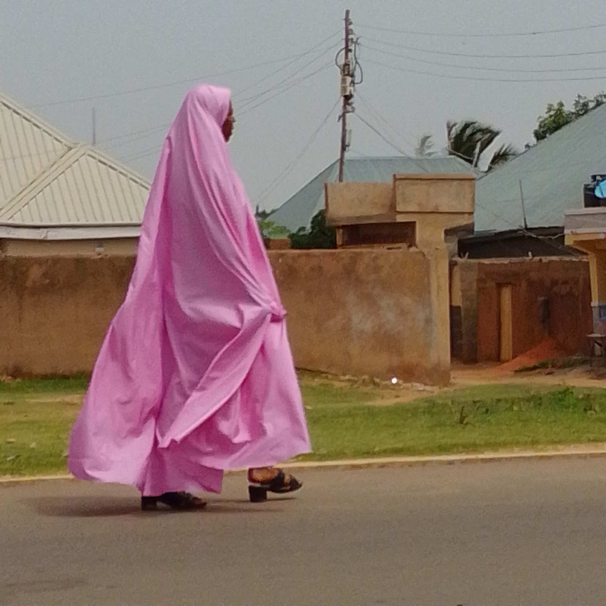 An individual, likely female, is depicted from the chest down, walking on an asphalt road. They are wearing a flowing, bright pink garment that covers their entire body except for their feet and lower legs. The garment has significant draping and movement, suggesting it is lightweight fabric. The individual is wearing dark, open-toed sandals. The background consists of a dirt path, a strip of green grass, and various buildings constructed with earth-toned walls. In the distance, there are corrugated metal roofs and a utility pole with multiple wires. The sky is overcast and light gray. The scene is outdoors, likely in a residential or semi-urban area.