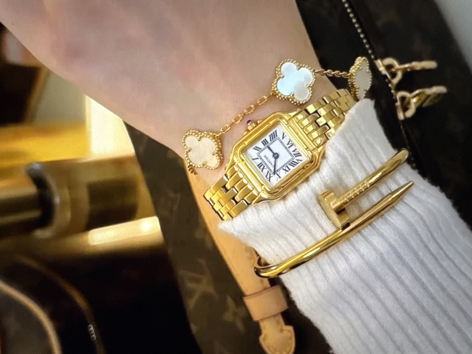 The image shows a close-up of a person's left wrist and forearm, adorned with several pieces of jewelry. The most prominent items are a gold wristwatch with a white face and Roman numerals, and a gold bracelet featuring several clover-shaped charms with mother-of-pearl inlays. A thick gold bangle, designed to resemble a nail, is also worn on the wrist. The background is blurred but appears to include a textured surface with a repeating pattern, possibly a handbag or wallet, and a hint of a reflective golden object. The overall impression is one of luxury and personal adornment. The location is not specified in the image.