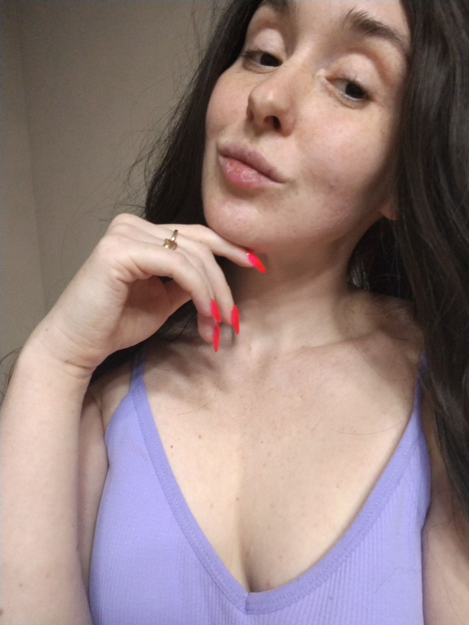 A woman with fair skin and freckles poses indoors. She is wearing a light purple ribbed tank top. Her dark brown hair is wavy and falls to her shoulders. She has a gold ring on her finger, and her nails are painted bright red. Her right hand is raised to her chin, and she is looking at the camera with a slight smile. The background is a plain light beige wall. There is no indication this image was taken in Izhevsk, Russia.