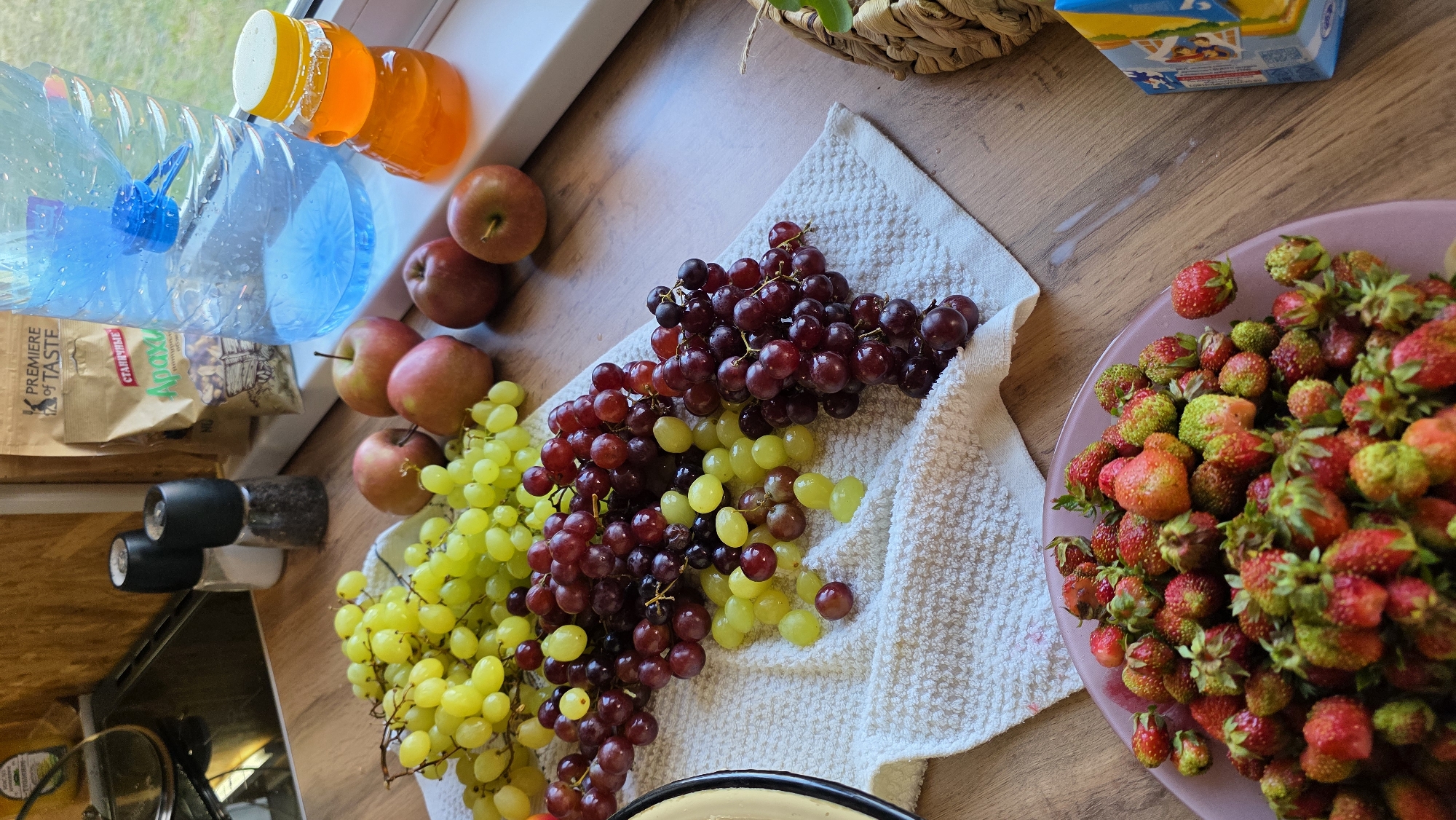 The image shows a collection of fruits and pantry items on a wooden countertop in what appears to be a kitchen. In the foreground, a pink bowl is filled with ripe strawberries. Behind the bowl, on a white towel, are several bunches of grapes, including green and red varieties. To the right of the grapes, there are a few red apples. Further back, on a white windowsill, sits a large blue plastic bottle of water, a small glass jar of honey, and a bag of peanuts. Two salt and pepper grinders are visible on the countertop as well. The background reveals a portion of a stove and a cabinet. The overall scene suggests a domestic setting, likely a kitchen, in the city of Kup'yarovo, Russia.