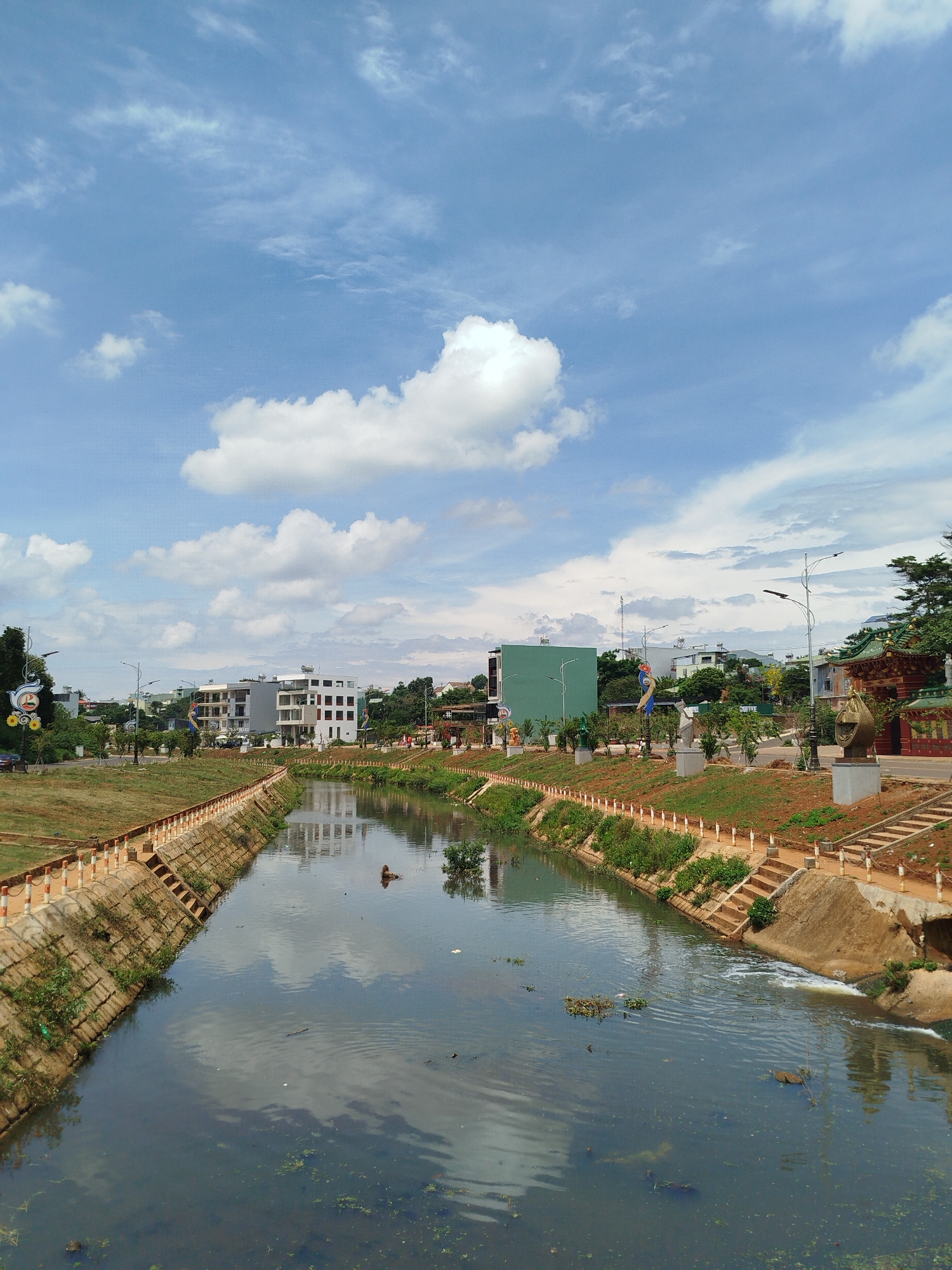 A long drainage canal traverses Pleiku Ward, Vietnam, with concrete embankments on either side. Vegetation grows along the edges. The water is murky with visible algae and debris. A light-colored fence runs along the edge of the embankment. In the distance, buildings and street lights line the banks. The sky is blue with scattered clouds. The architecture reflects a mix of modern and traditional Vietnamese styles. A set of concrete stairs leads down to the canal's edge.