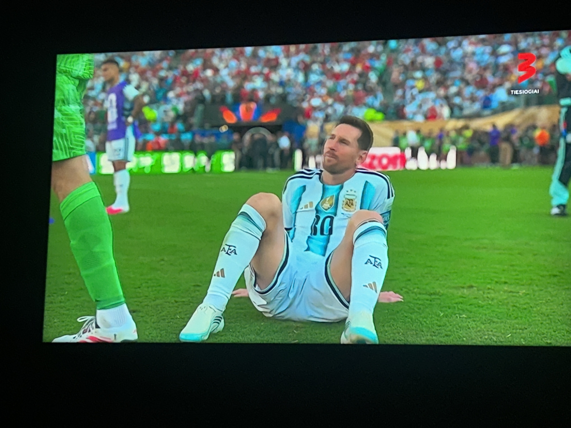 In the city of Unknown, Unknown, a soccer player in an Argentina jersey and shorts is sitting on the grass on a soccer field. He is looking up and to his right. There are other players and a crowd in the background. The scoreboard in the background shows a "3".