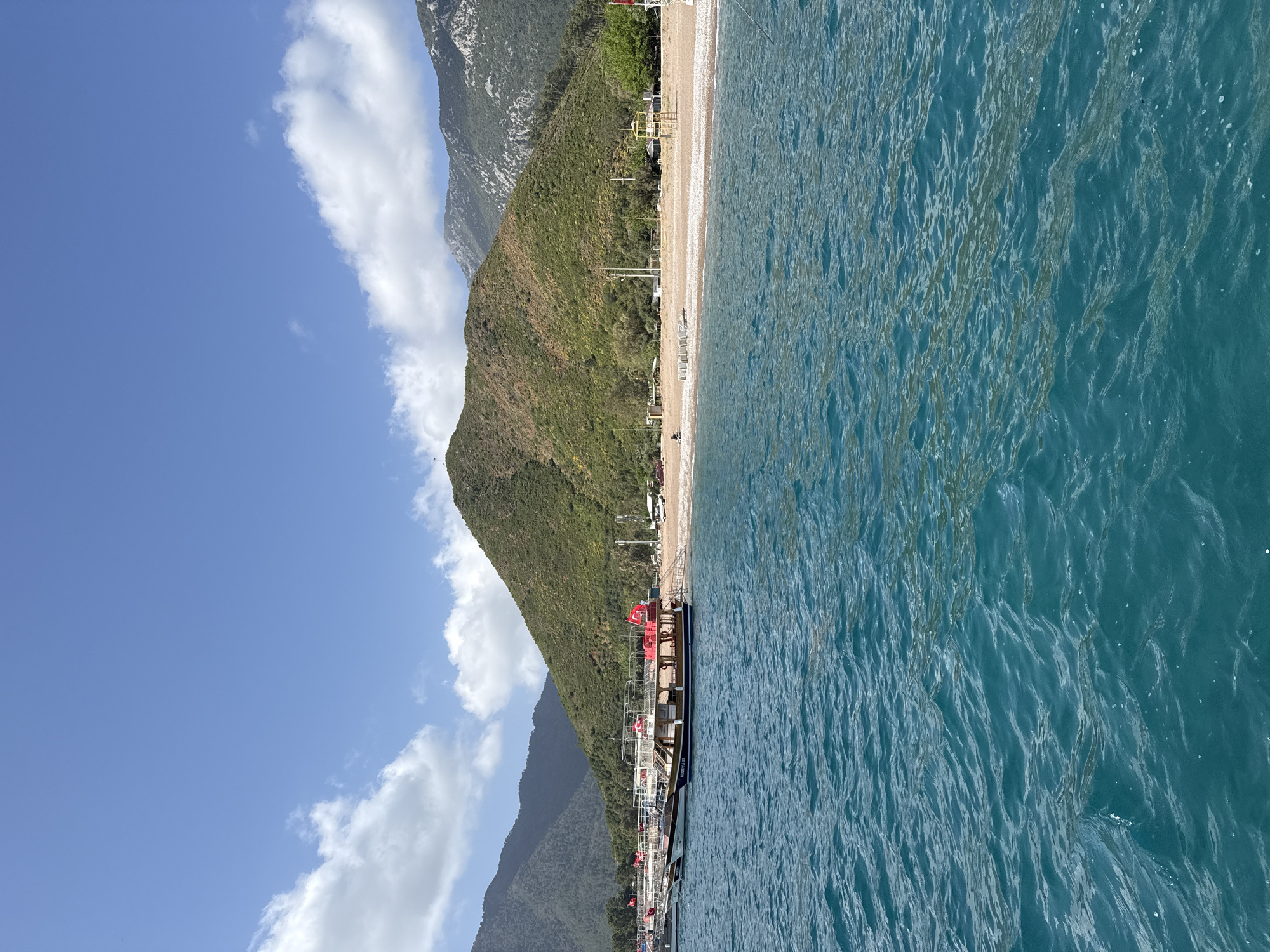 The image depicts a coastal scene in Kumluca, Turkey, featuring a beach bordered by a mountain range on one side and the Mediterranean Sea on the other.
The beach is light-colored and appears to be composed of sand or small pebbles. A few structures, possibly beach huts or buildings, are visible along the coastline.
The mountain range rises steeply behind the beach, covered in green vegetation. The sky is blue with scattered white clouds. The sea is calm with a deep blue color.
A long pier runs along the beach. It appears to accommodate several large boats or yachts. Red flags are visible on the vessels.