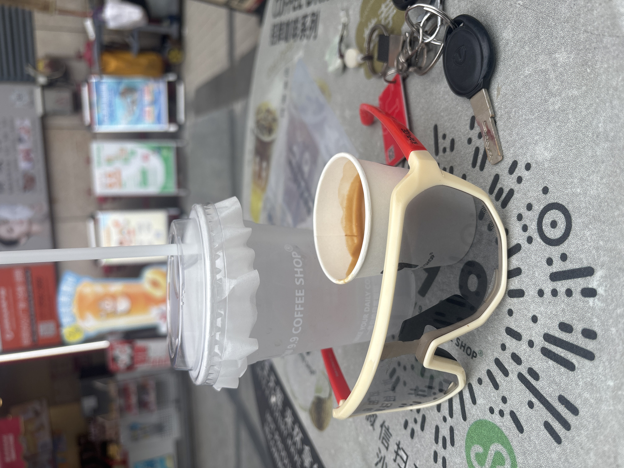 A close-up shot focuses on a table outdoors, likely at a coffee shop in Nanhai District, China, given the presence of Chinese text in the background. On the table, a large plastic cup with a paper sleeve and a straw sits to the left. The cup is from "M9.9 COFFEE SHOP". To the right of the cup is a small paper cup filled with espresso. In the foreground, a pair of large, cream-colored sunglasses with red accents is placed diagonally. A set of keys, including a car key with a black fob, is to the right of the sunglasses. The table surface appears to be covered with a patterned mat, possibly featuring QR codes. In the blurred background, signs and building facades suggest an urban commercial setting.