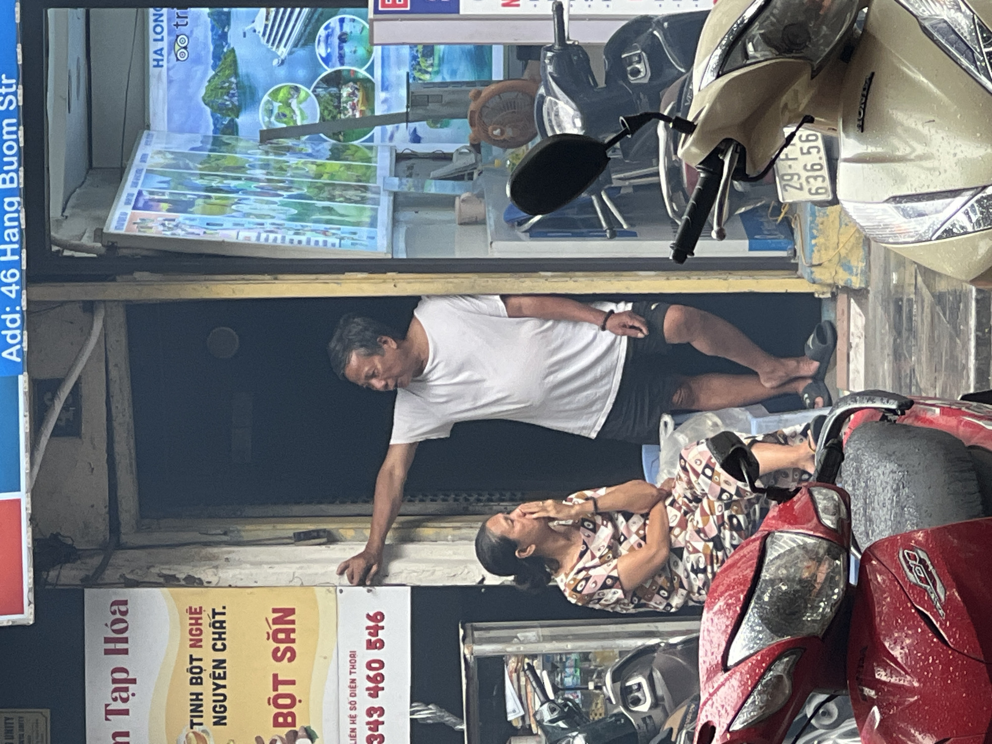 The image captures a street scene in Hanoi, Vietnam, likely in the Old Quarter given the style of the buildings and shop fronts. The focal point is an open doorway of a small shop selling what appears to be food products, indicated by signage in Vietnamese including "TINH BỘT NGHỆ NGUYÊN CHẤT" (Pure Turmeric Starch) and "BỘT SẮN" (Cassava Flour). A man in a white t-shirt and black shorts is leaning against the doorframe, looking to his right. A woman, seated on a blue plastic chair to the left, is wearing a patterned dress and has her hand to her face, appearing to be wiping away tears or resting her chin. Several motorbikes are parked in front of the shop, some of them in the foreground. A large poster advertising tours to Ha Long Bay is visible inside the shop. The ground is wet, suggesting recent rain.