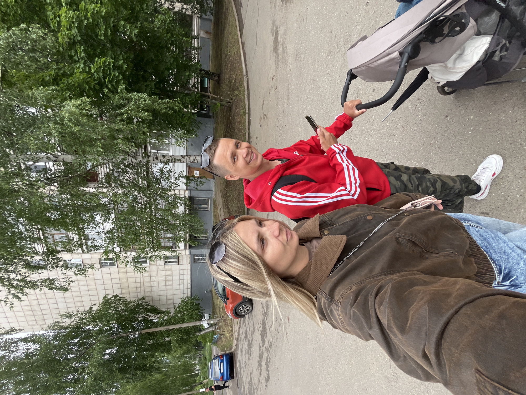 A couple poses for a selfie on a city street. The woman, with blonde hair and sunglasses perched atop her head, wears a brown jacket and blue jeans. The man, beside her, wears a red Adidas tracksuit jacket with white stripes and camouflage pants, and he holds a smartphone. He also has sunglasses on his head and pushes a baby stroller. In the background, there are apartment buildings, green trees, and parked cars, including a red car and a blue car. This image was taken in the city of Chaikovsky, Russia.