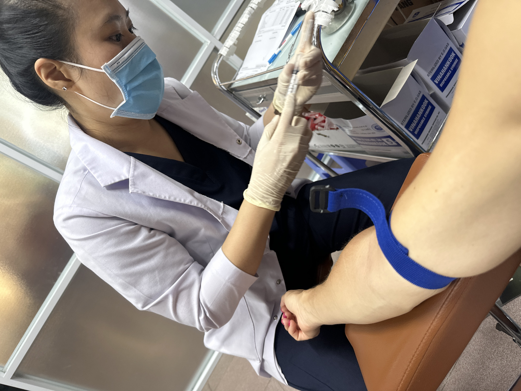 A close-up, high-angle shot shows a medical professional in Nha Trang Ward, Vietnam, preparing to draw blood from a patient's arm. The medical professional, wearing a light blue surgical mask and pale yellow latex gloves, holds a syringe with a needle. The patient's arm, resting on a brown padded chair, has a blue tourniquet applied above the injection site. The background features a metal medical cart with medical supplies, including boxes labeled "VINAHANKOOK DISPOSABLE SYRINGE." The overall environment appears to be a clinical setting.