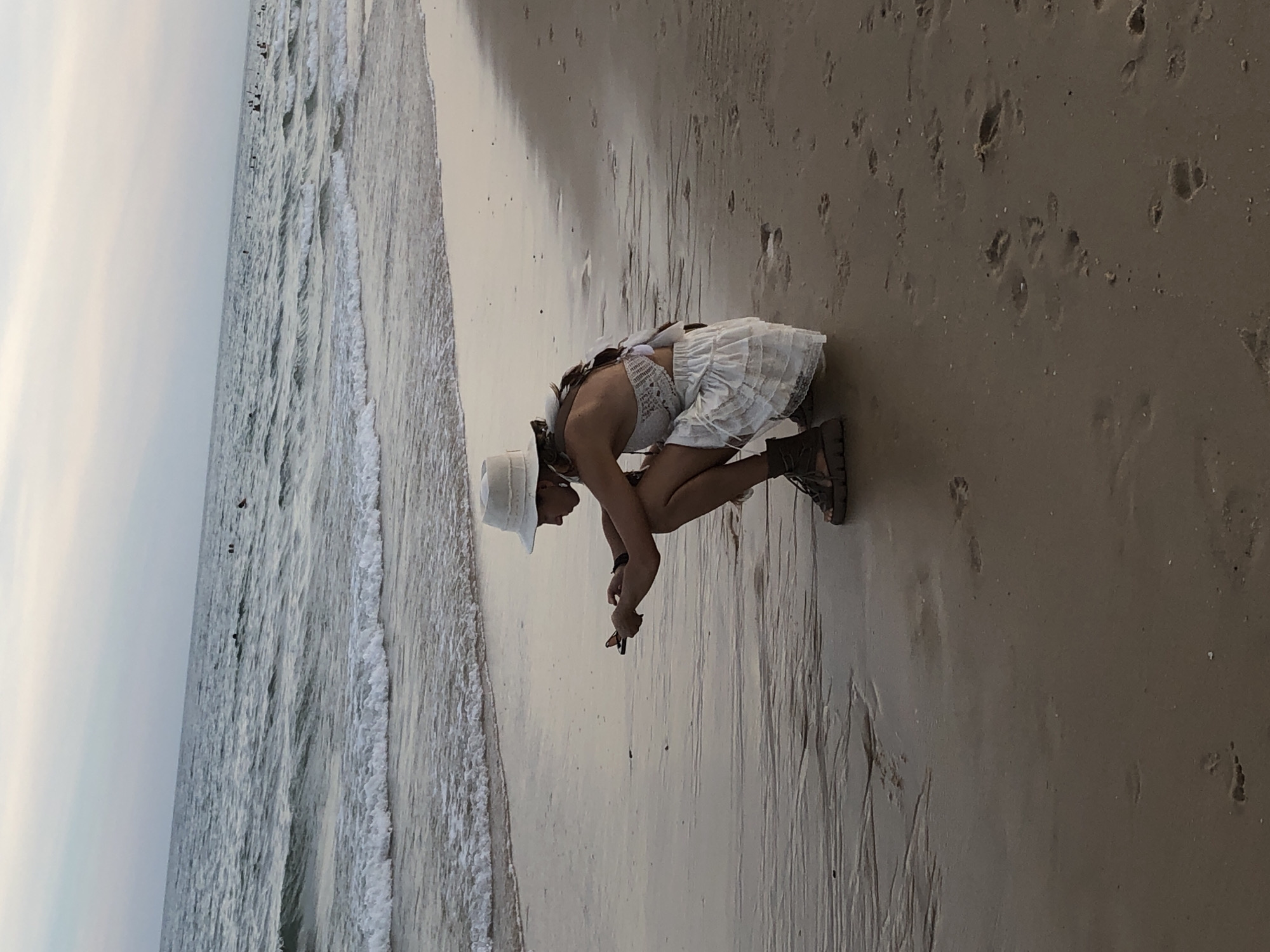 In this image, a person is seen crouching on a sandy beach with the ocean to their left. The person is wearing a white hat, a white top with a textured pattern, and a white skirt. They are also wearing brown sandals. Their posture suggests they are examining something on the sand. The water is calm with small waves lapping at the shore. The sky is overcast, indicating a cloudy day. There are numerous footprints visible in the wet sand. The scene appears to be set in the late afternoon or early evening. The location is identified as Phu Thuy Ward, Vietnam. There is no visible text in the image.